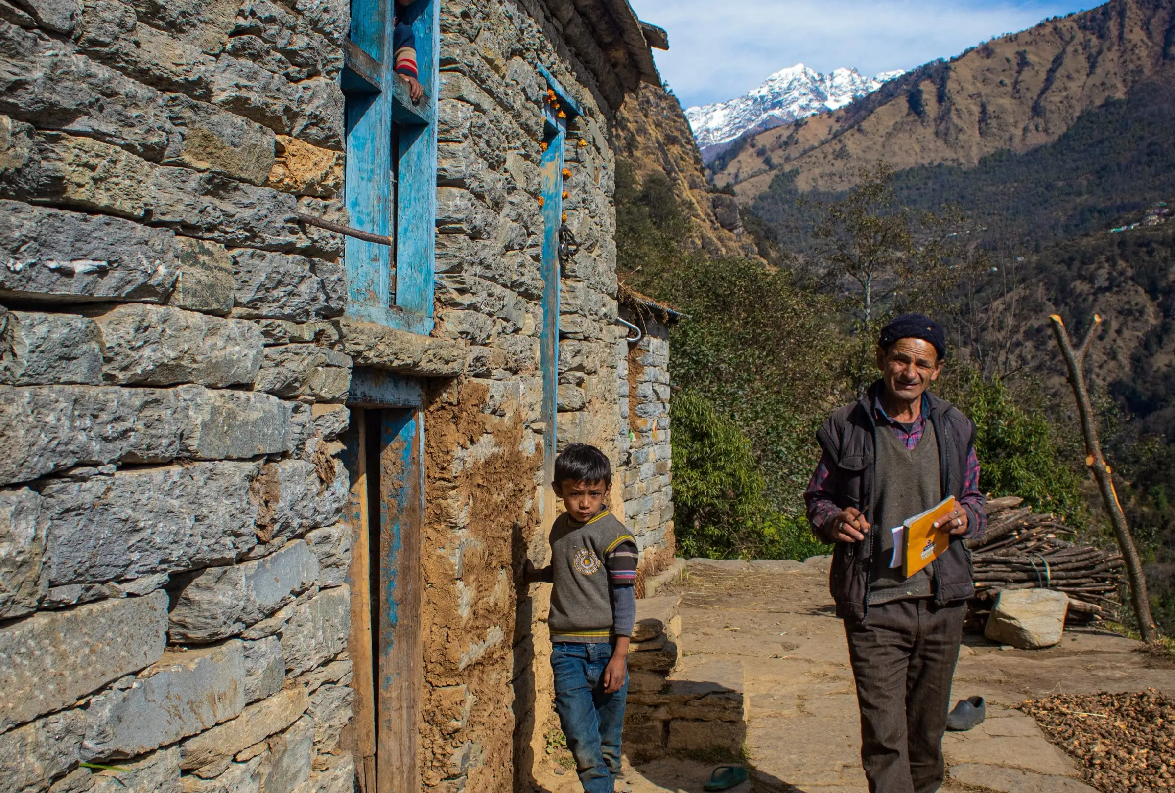 A villager and a boy outside a stone house with blue windows in Namik village, with forested slopes and a snow peak behind