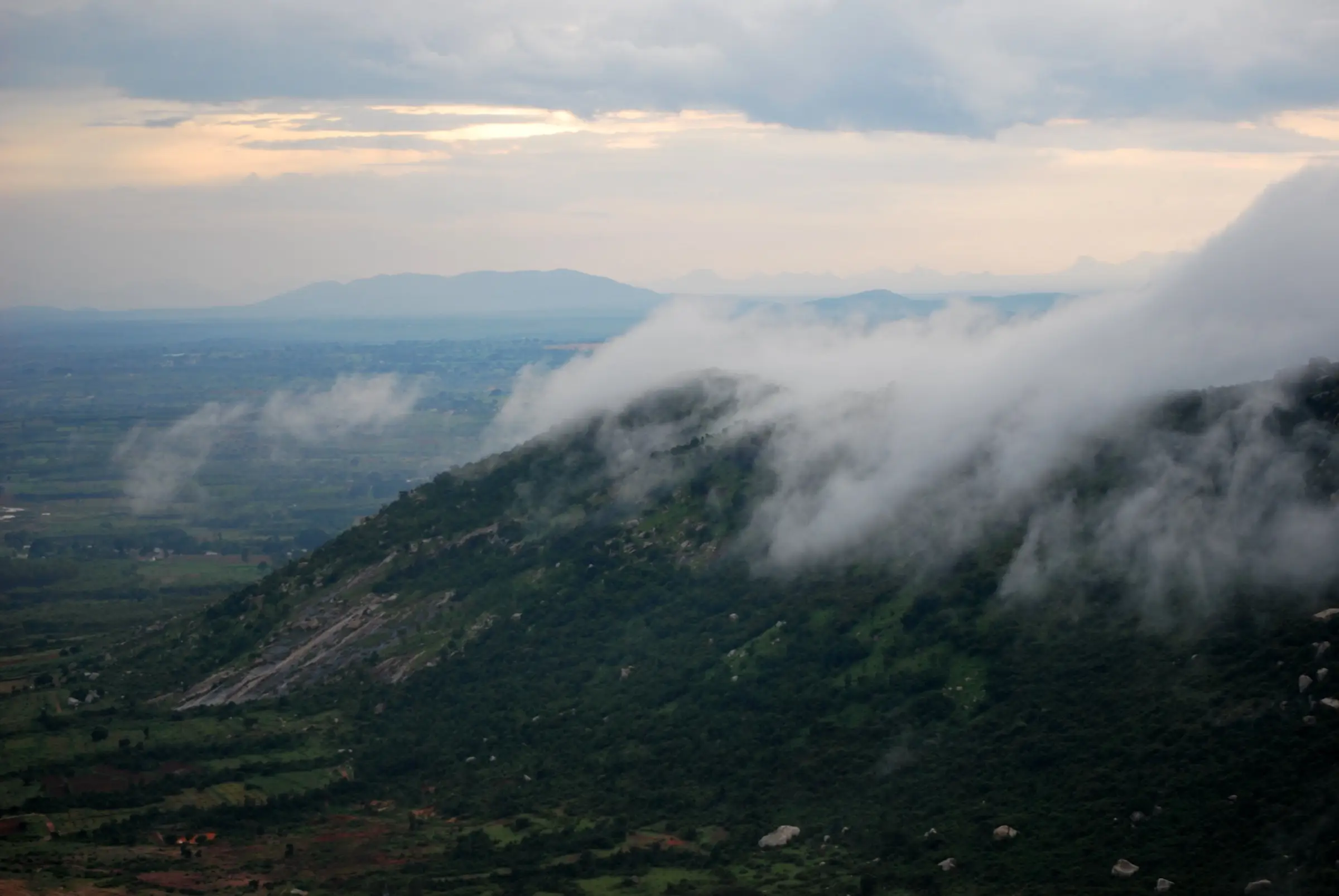 Monsoon clouds rolling over a forested hillside of the Nandi Hills range above farmland, near the Dibbagiri trek