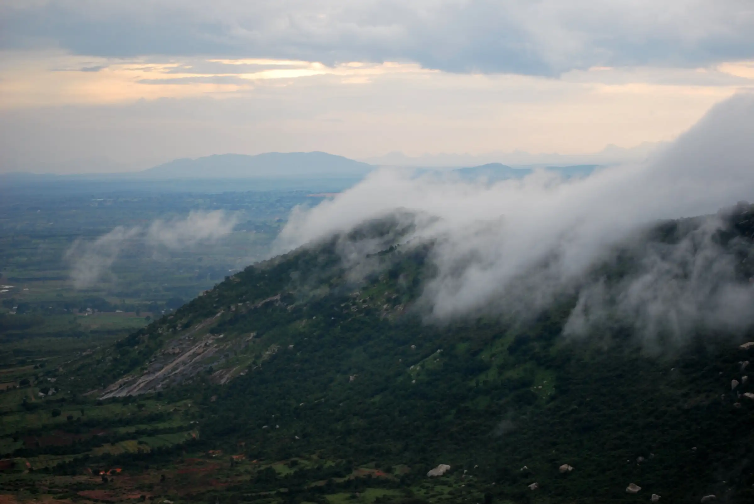 Monsoon clouds rolling over a forested hillside of the Nandi Hills range above farmland, near the Dibbagiri trek