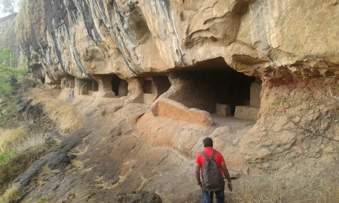 A trekker with a backpack walks along the dry rock ledge towards the Thanale cave openings under the cliff