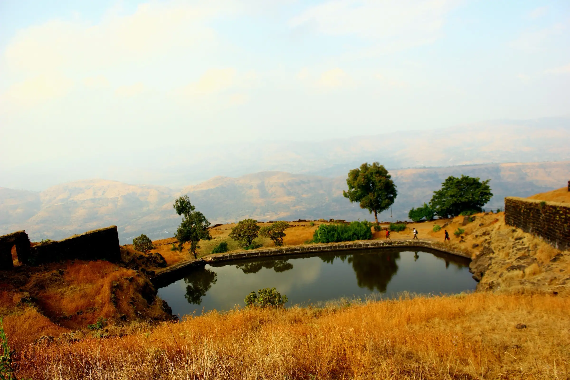 Padmavati lake inside Rajgad Fort with old walls, trees and walkers, and hazy Sahyadri hills behind