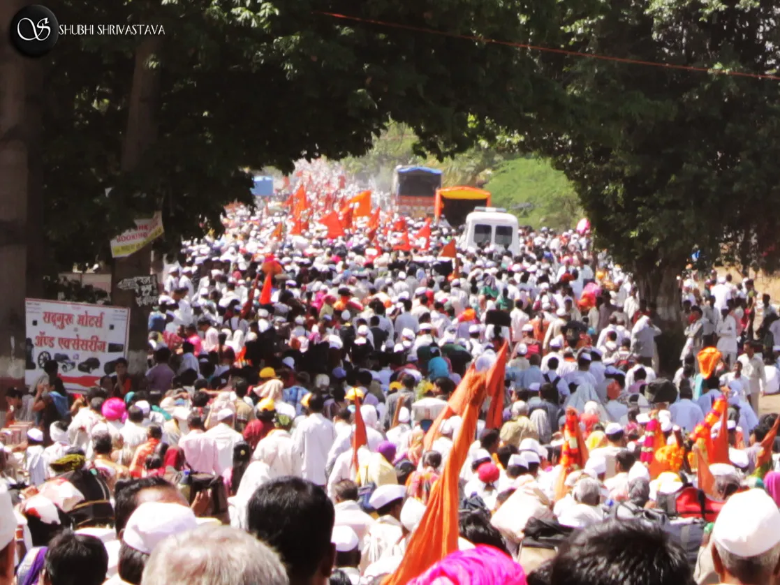 Thousands of warkaris in white caps with saffron flags walking along a tree-lined road with the palkhi procession near Pune