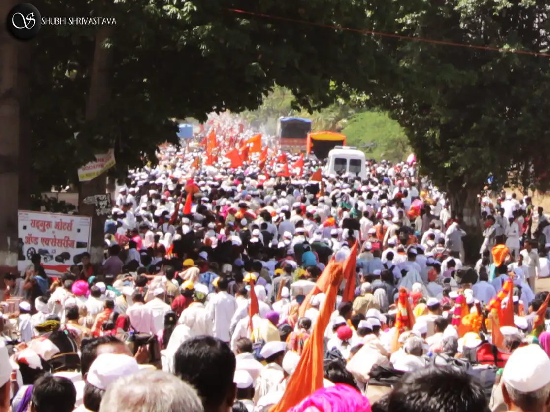 Thousands of warkaris in white caps with saffron flags walking along a tree-lined road with the palkhi procession near Pune