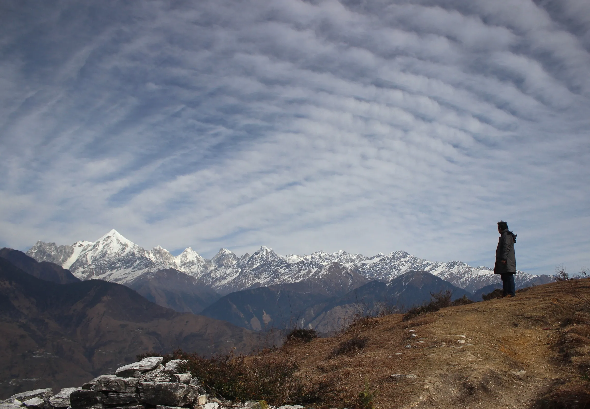 A trekker on a grassy ridge at Munsiyari, the start of the Ralam Glacier trek, facing the snow-covered Panchachuli range under rippled cloud