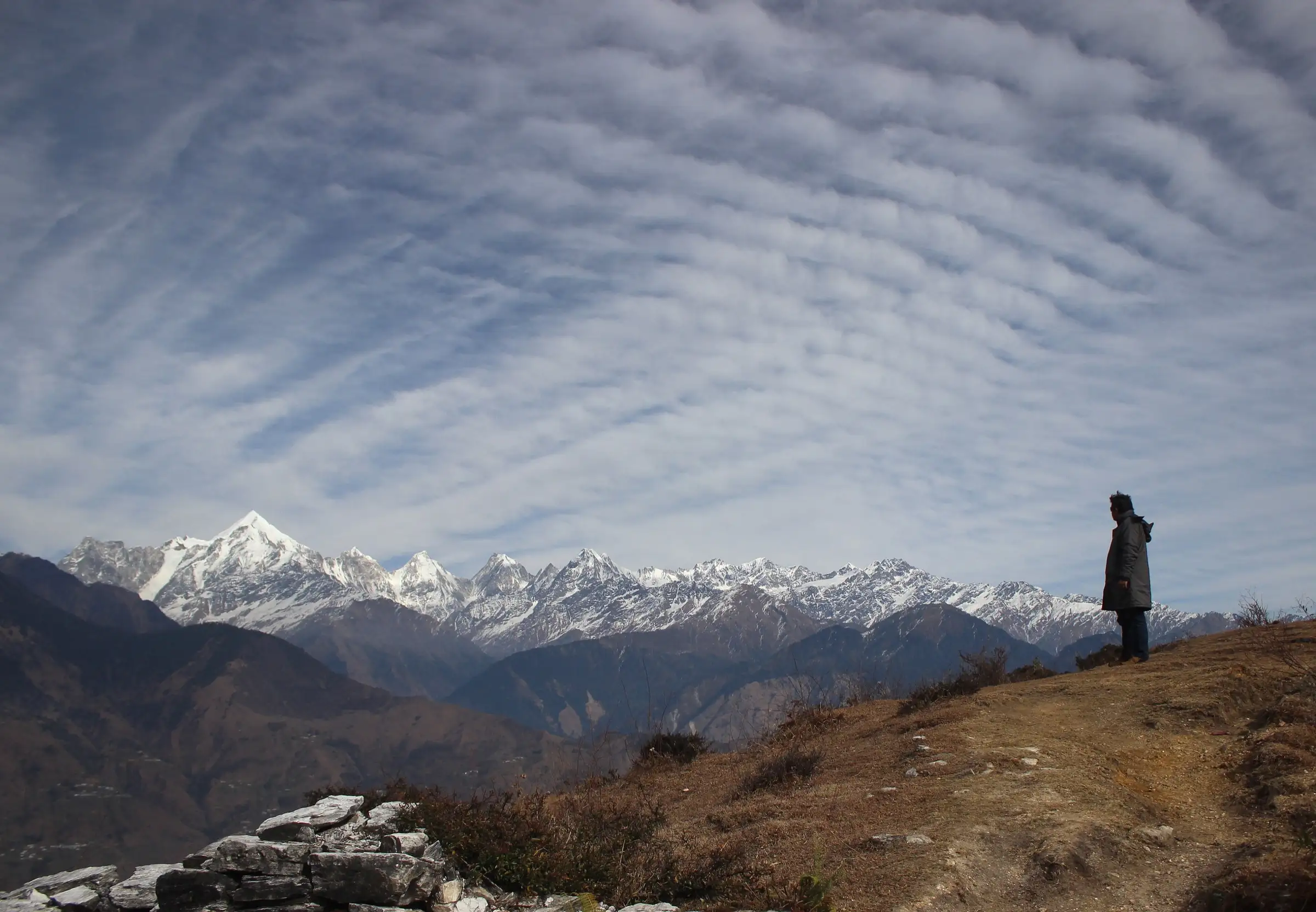 A trekker on a grassy ridge at Munsiyari, the start of the Ralam Glacier trek, facing the snow-covered Panchachuli range under rippled cloud