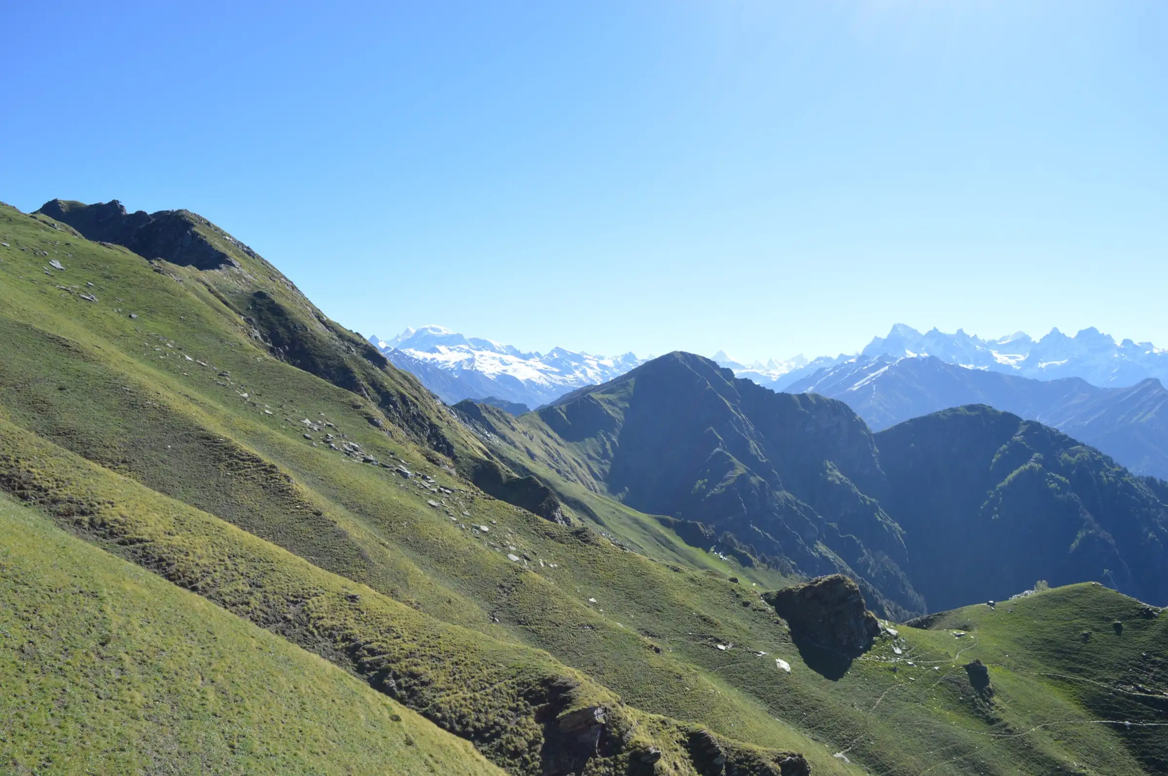 Green grassy ridge of Chanderkhani Pass falling into a deep valley with snow peaks on the horizon under a clear blue sky