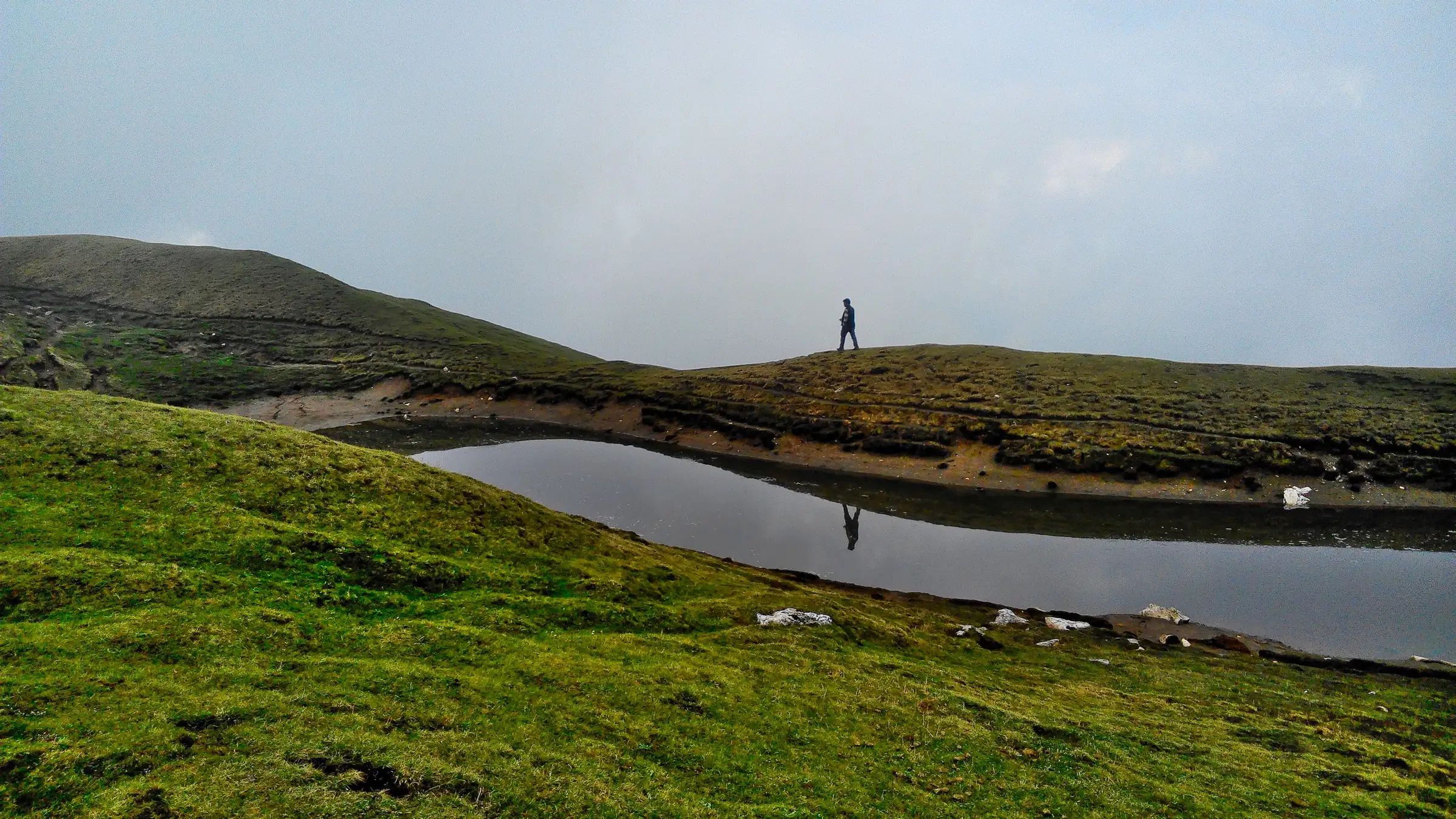 A trekker walking along a grassy ridge beside the small lake on top of Sar Pass, with mist all around