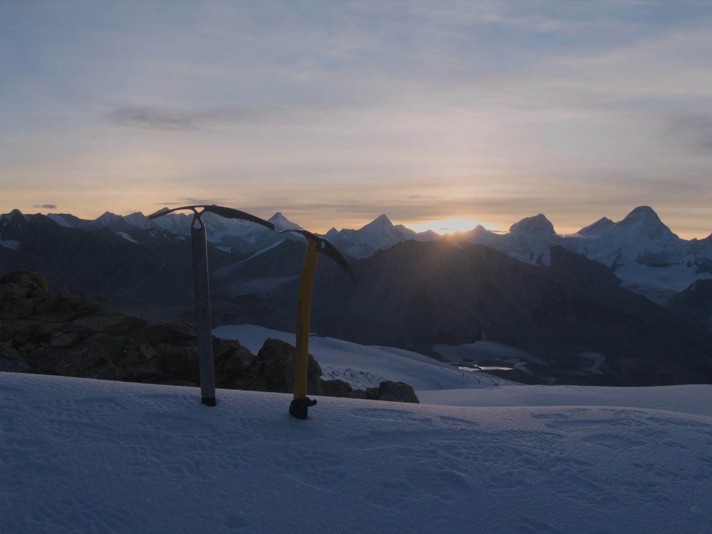 Two ice axes planted in snow on Kalindi Khal at sunrise, with snow peaks to the east on the horizon