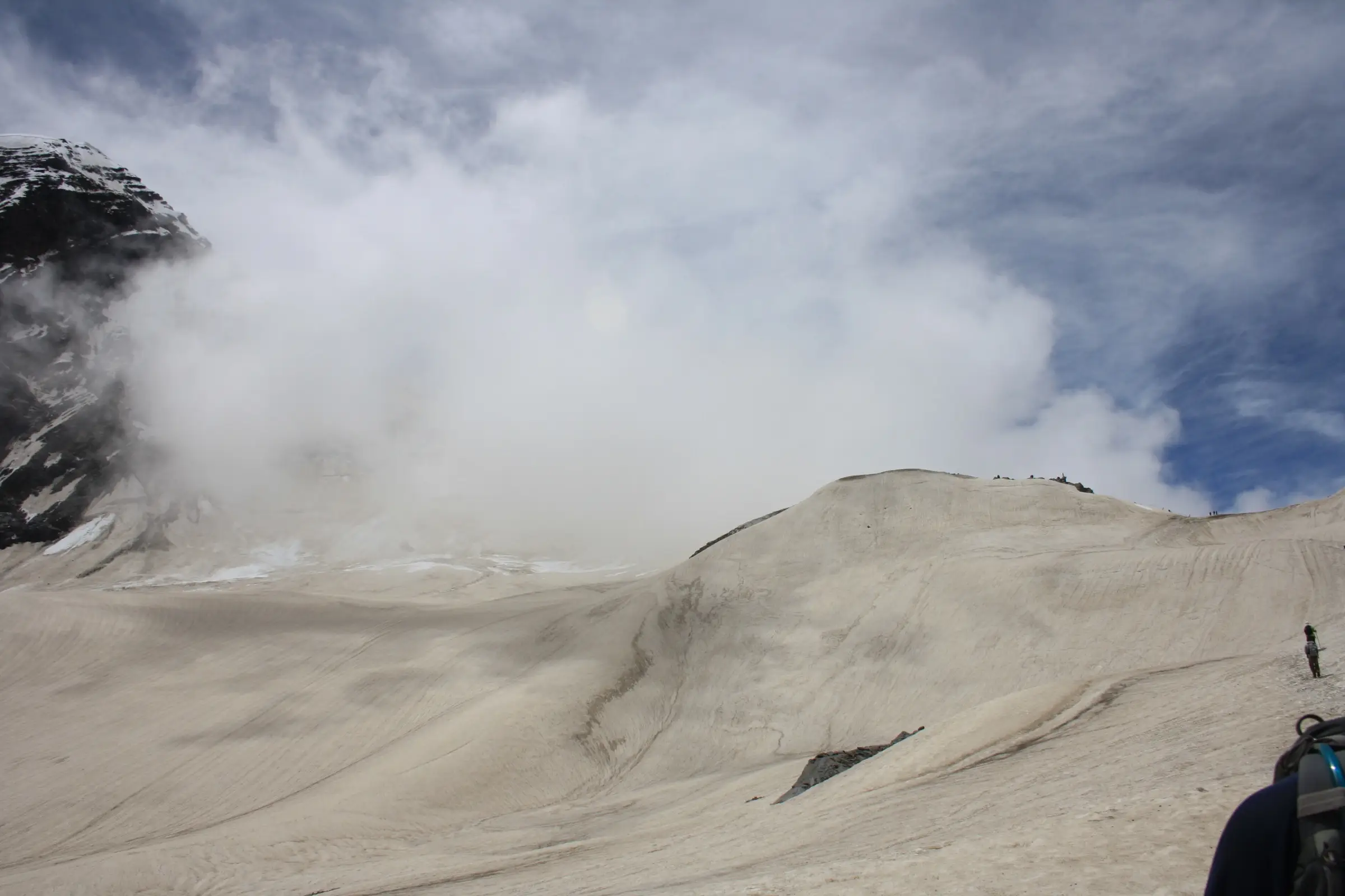 Snow-covered slope leading up to Pin Bhaba Pass with trekkers on the skyline and cloud rolling over a dark peak