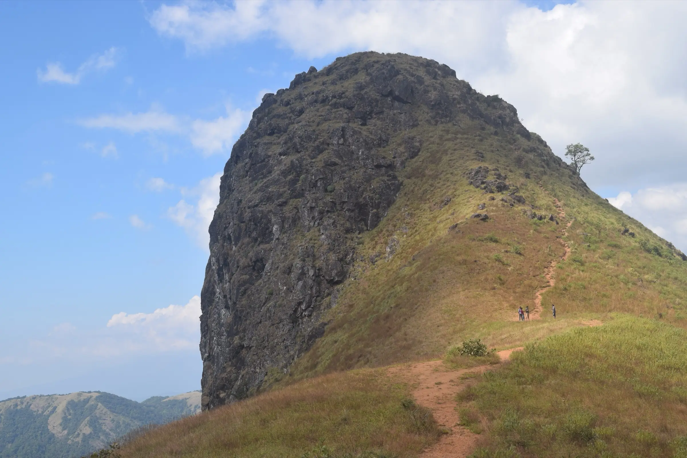 Close view of the steep rock face of Ettina Bhuja with trekkers on the grassy path below