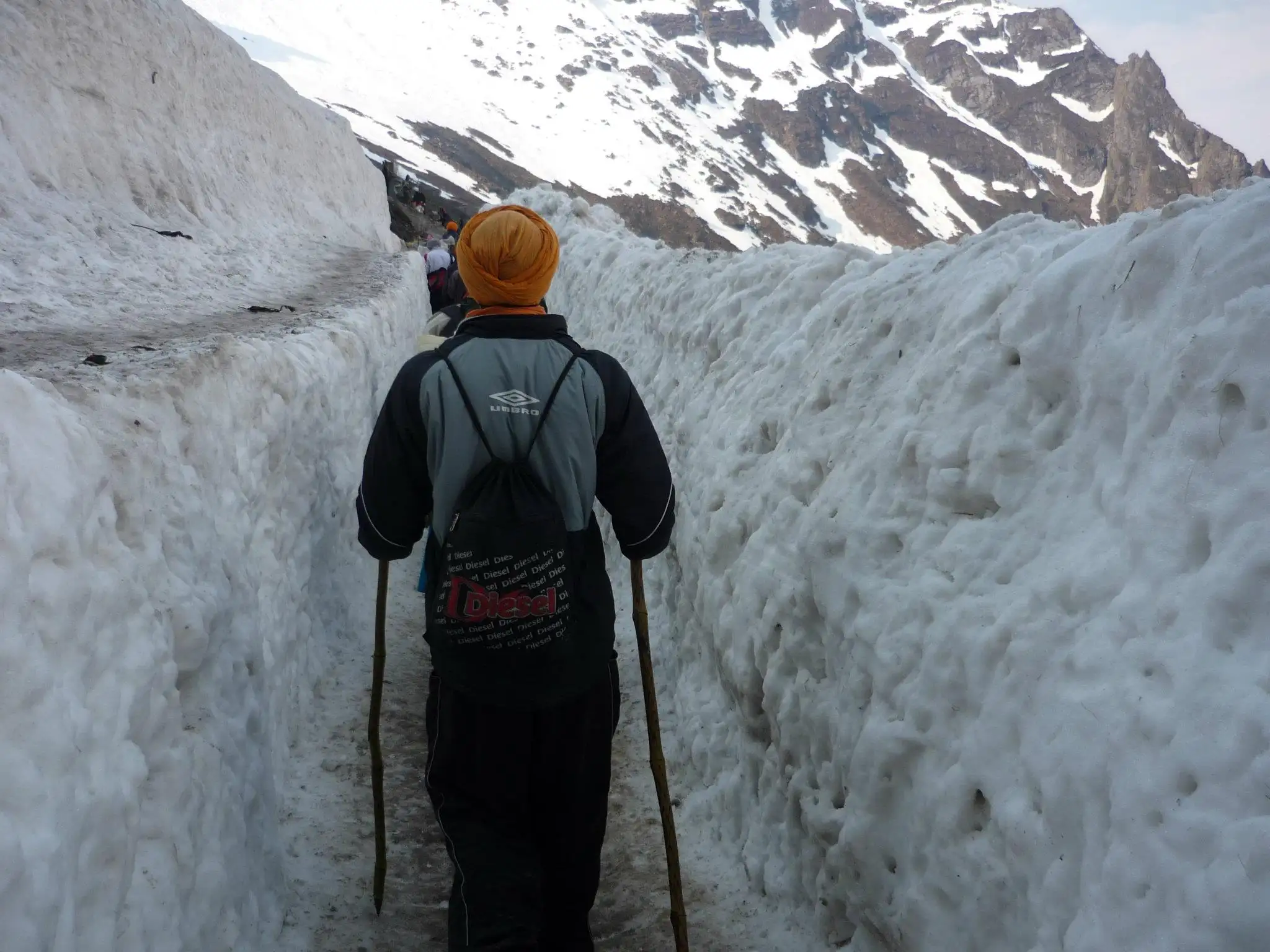Sikh pilgrim with two walking sticks on a path cut between high snow walls on the way to Hemkund Sahib
