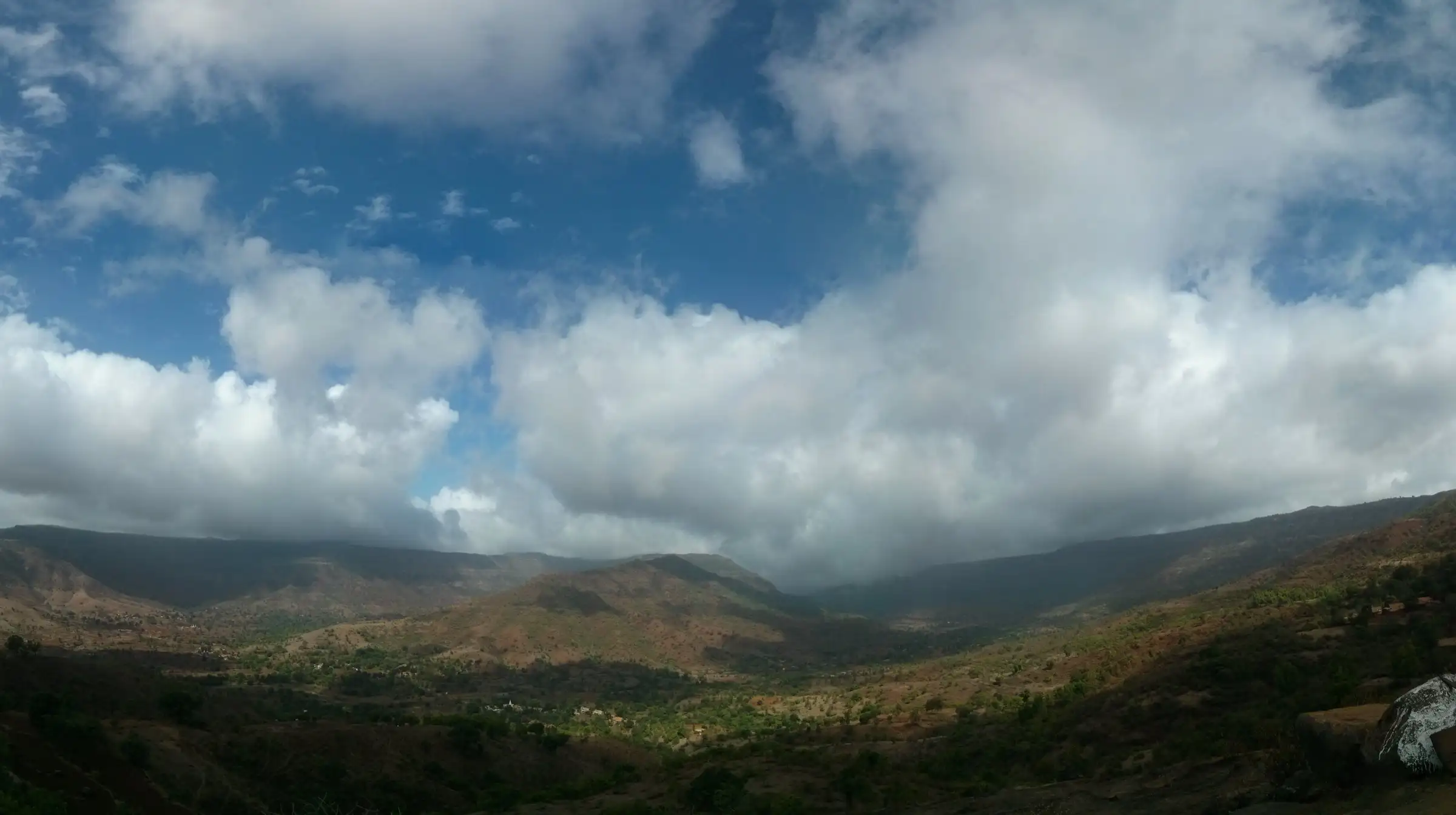Wide valley of green and red-earth hills below the Raireshwar plateau under heavy white monsoon clouds