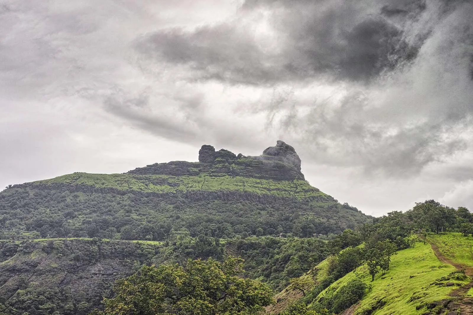 Irshalgad plateau and its twin rock pinnacle under grey monsoon clouds, with a grassy trail on a green ridge in the foreground