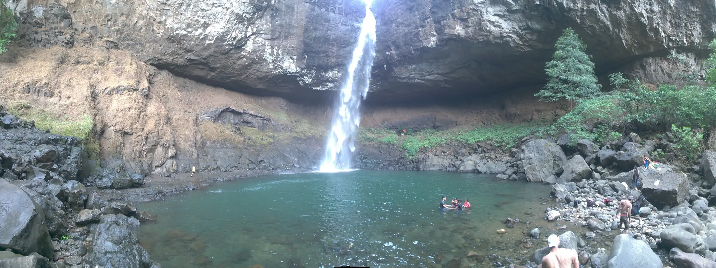 Panorama of the Devkund plunge pool ringed by boulders, with the waterfall pouring from an overhanging cliff