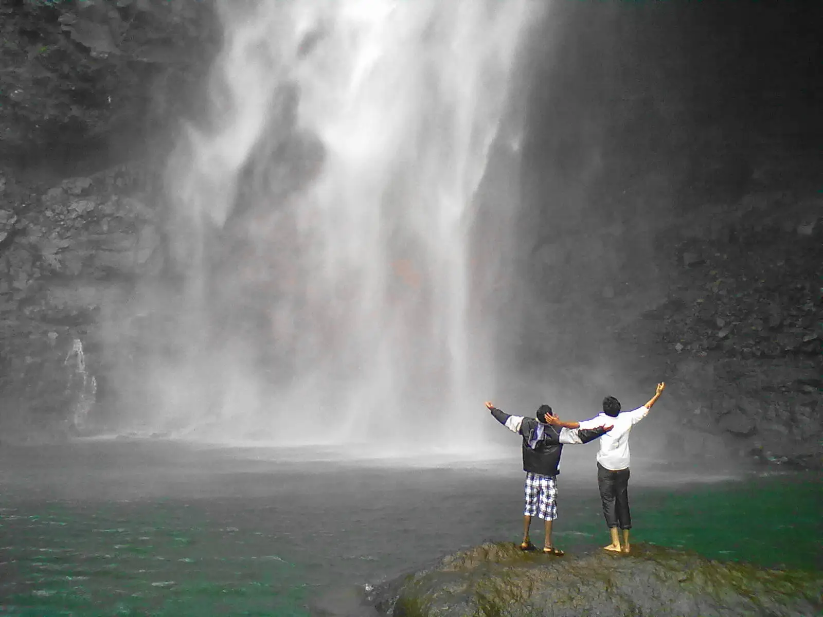 Two trekkers with arms raised on a rock beside the green plunge pool, facing the misty base of the waterfall