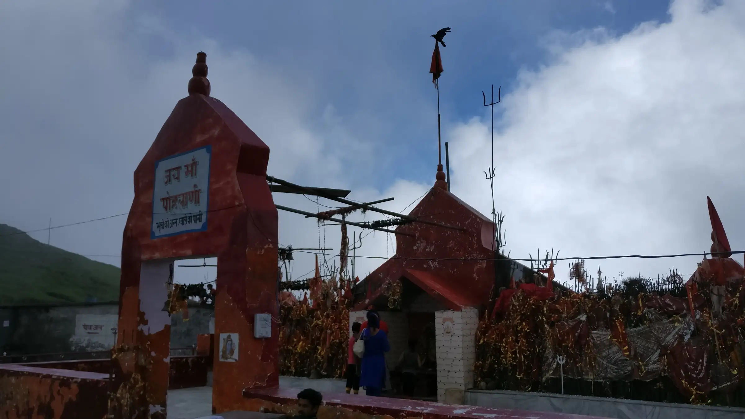 Red gateway reading Jai Shri Pohlani Mata and the small temple with trishuls and red chunris on Dainkund top