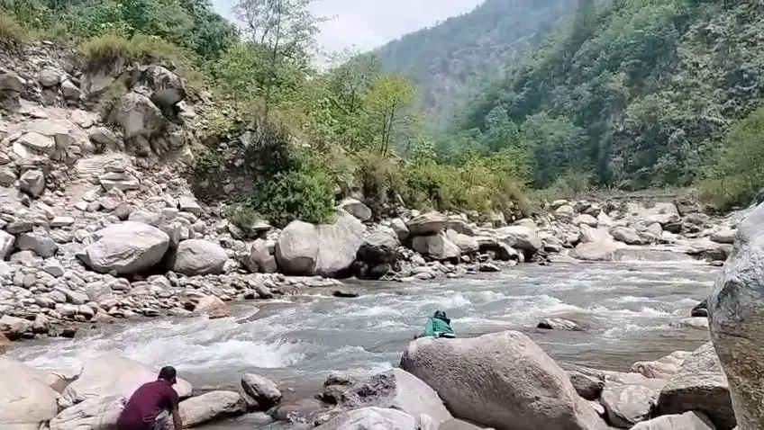 Trekkers resting on boulders beside the rushing Eastern Ramganga river in a forested valley below Namik