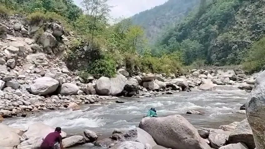 Trekkers resting on boulders beside the rushing Eastern Ramganga river in a forested valley below Namik