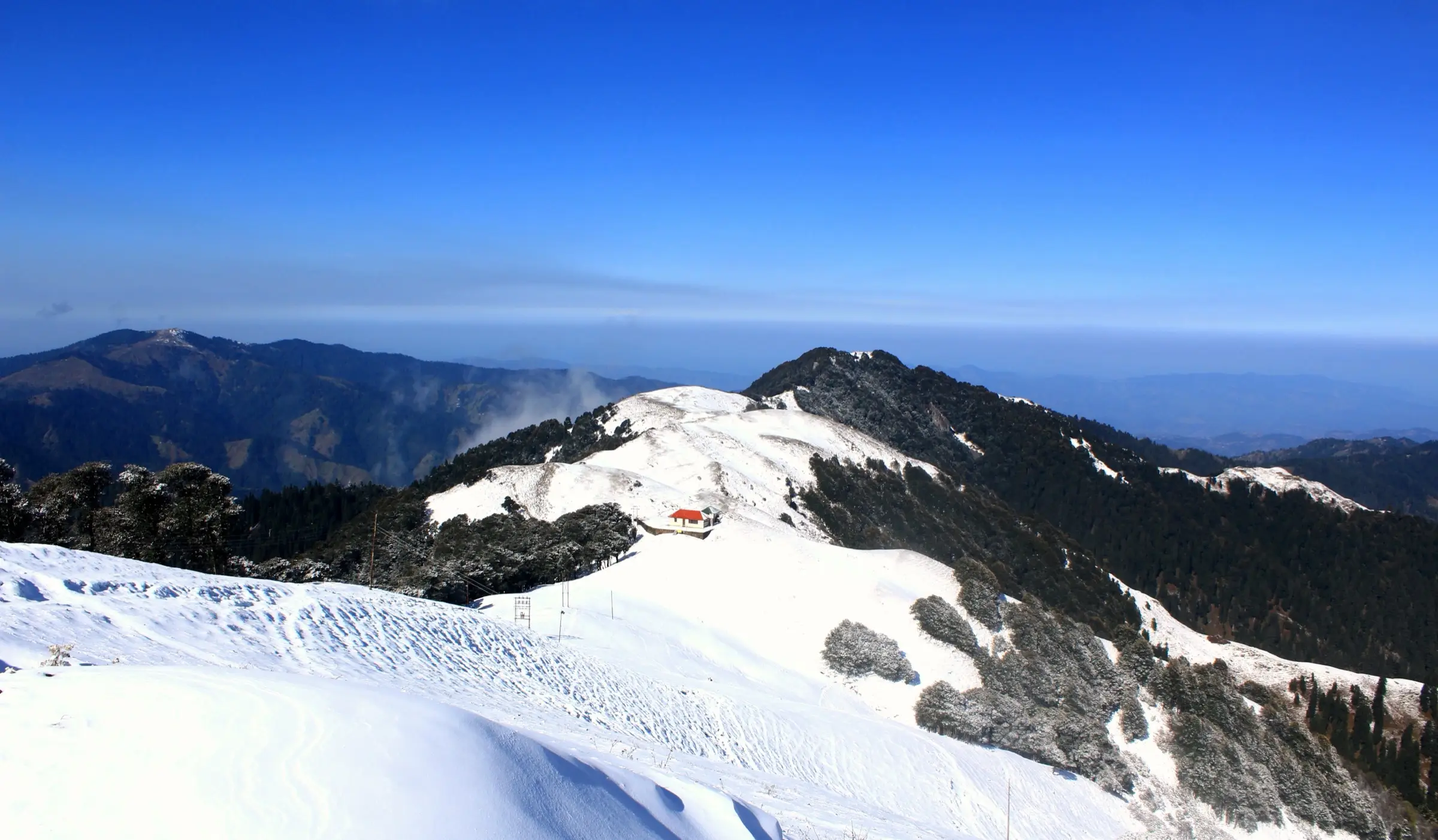 Snow-covered Shikari Devi ridge with a red-roofed building and snow-dusted forest under a clear blue sky
