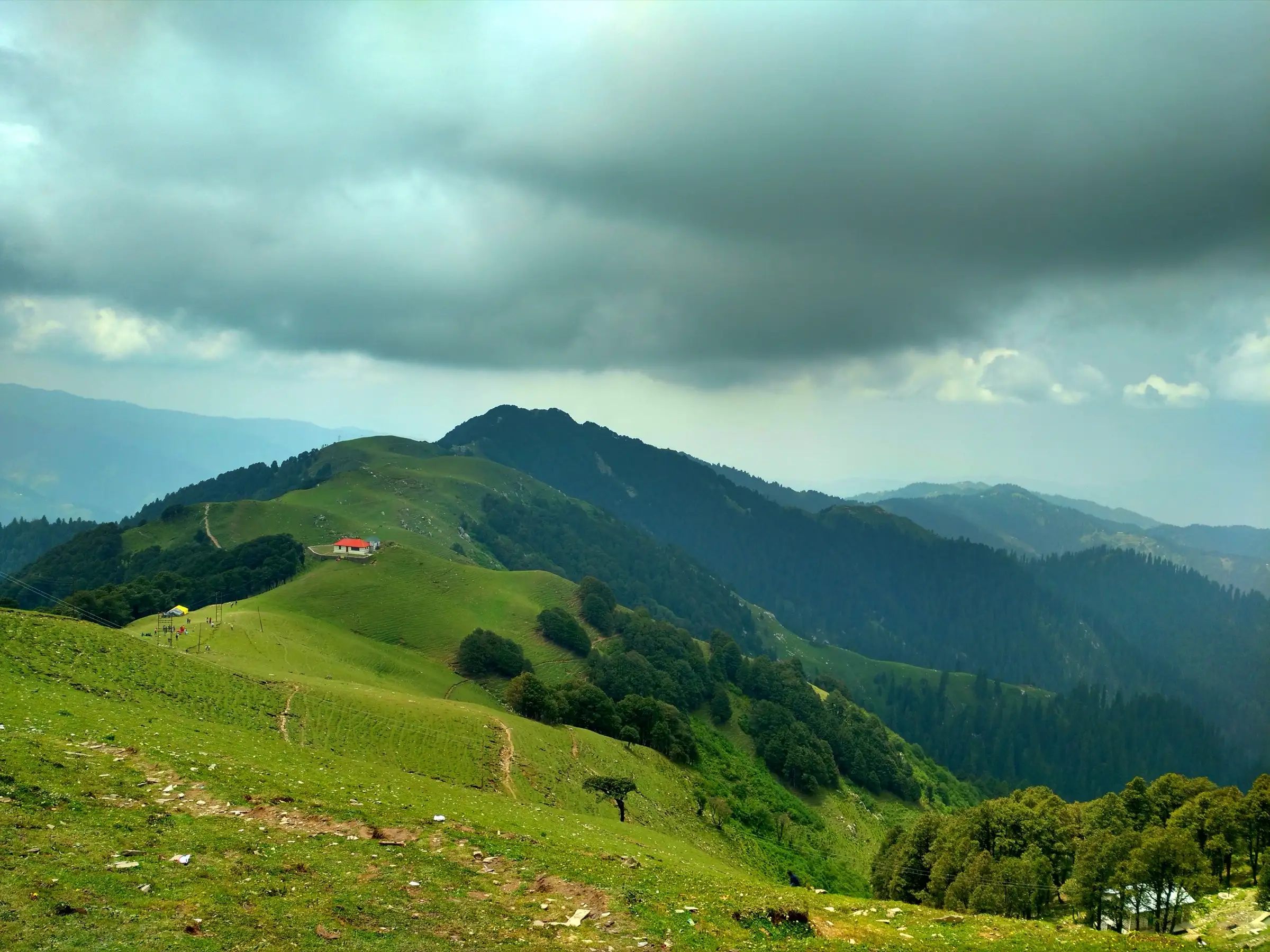 Green ridge-top meadows with a red-roofed hut and forested ridges under heavy monsoon cloud, on the Shikari Devi ridge above Janjehli