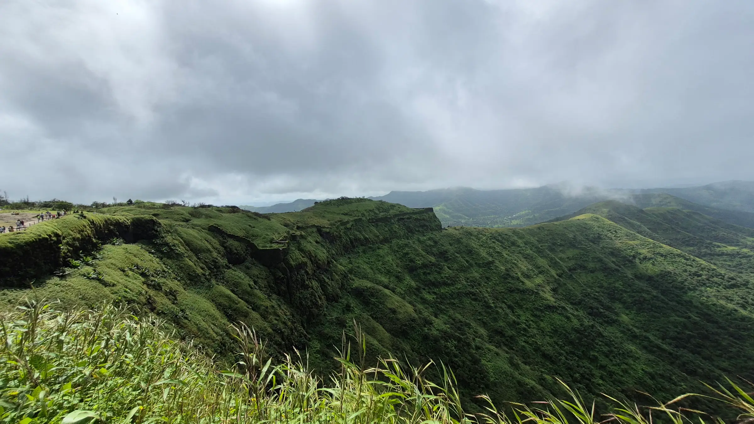 Grassy flat-topped ridge of Sinhagad Fort under grey monsoon clouds, with Sahyadri hills fading into the distance