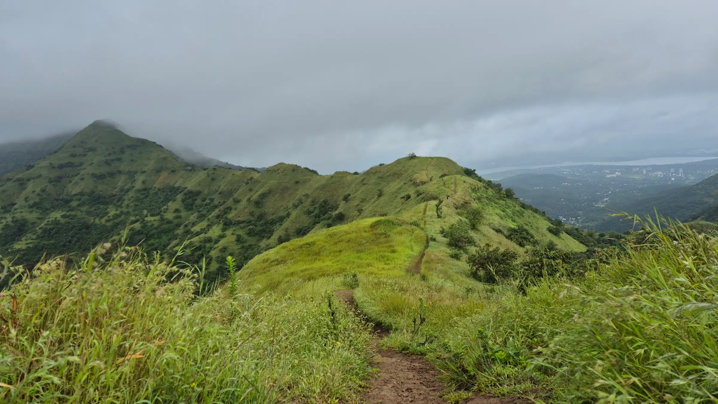 A narrow footpath running along a grassy ridge on Sinhagad towards a cloud-capped hilltop