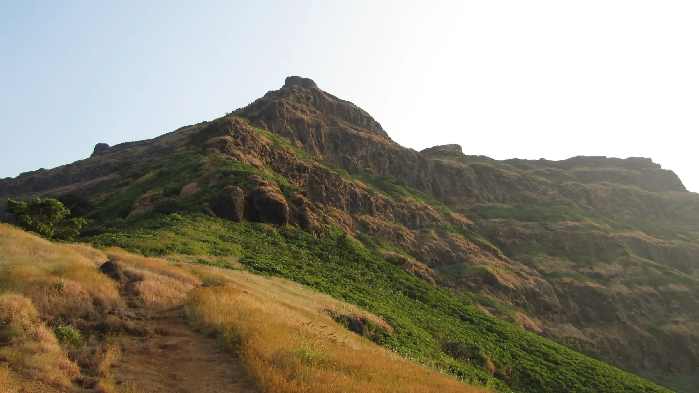 Dry grass trail on the ridge leading towards the rocky summit and cliffs of Torna Fort in clear weather