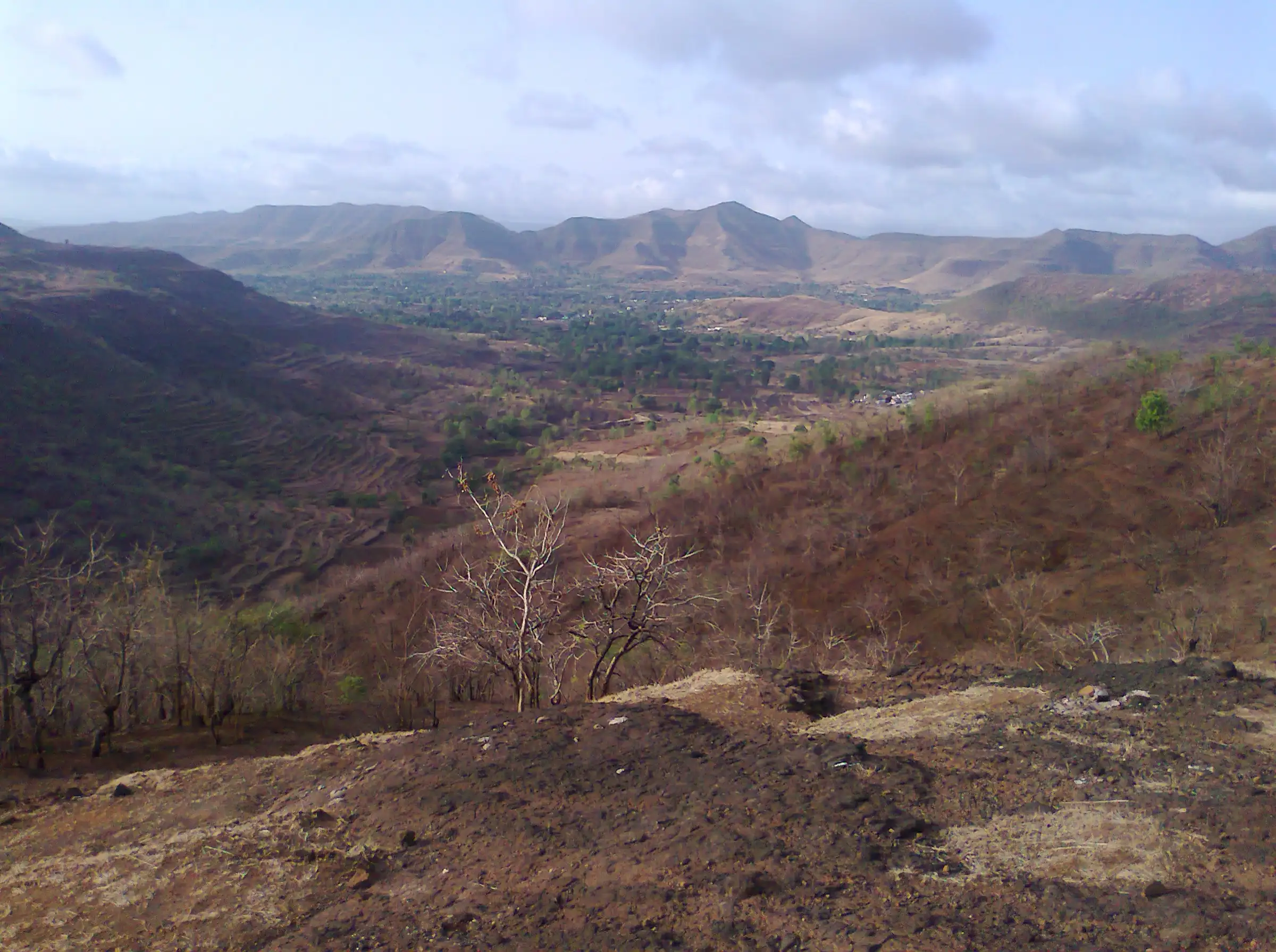 View from the Katraj-Sinhagad ridge over a green valley towards a line of Sahyadri hills, with dry grass and a bare earth slope in front
