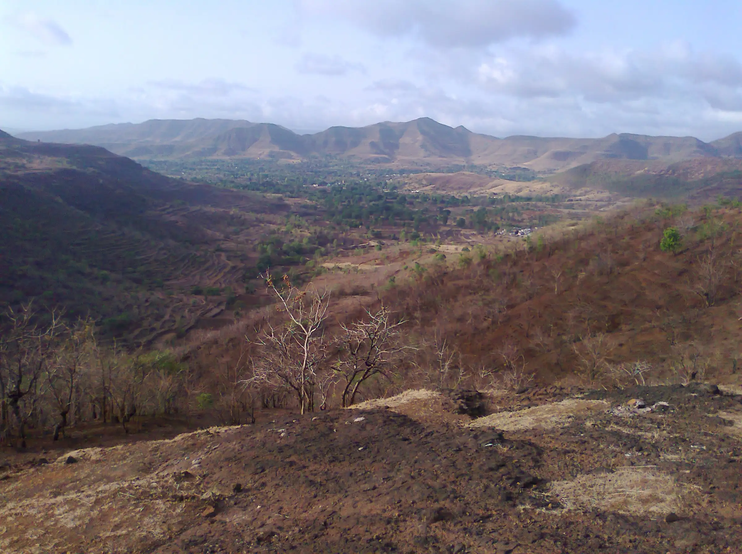 View from the Katraj-Sinhagad ridge over a green valley towards a line of Sahyadri hills, with dry grass and a bare earth slope in front