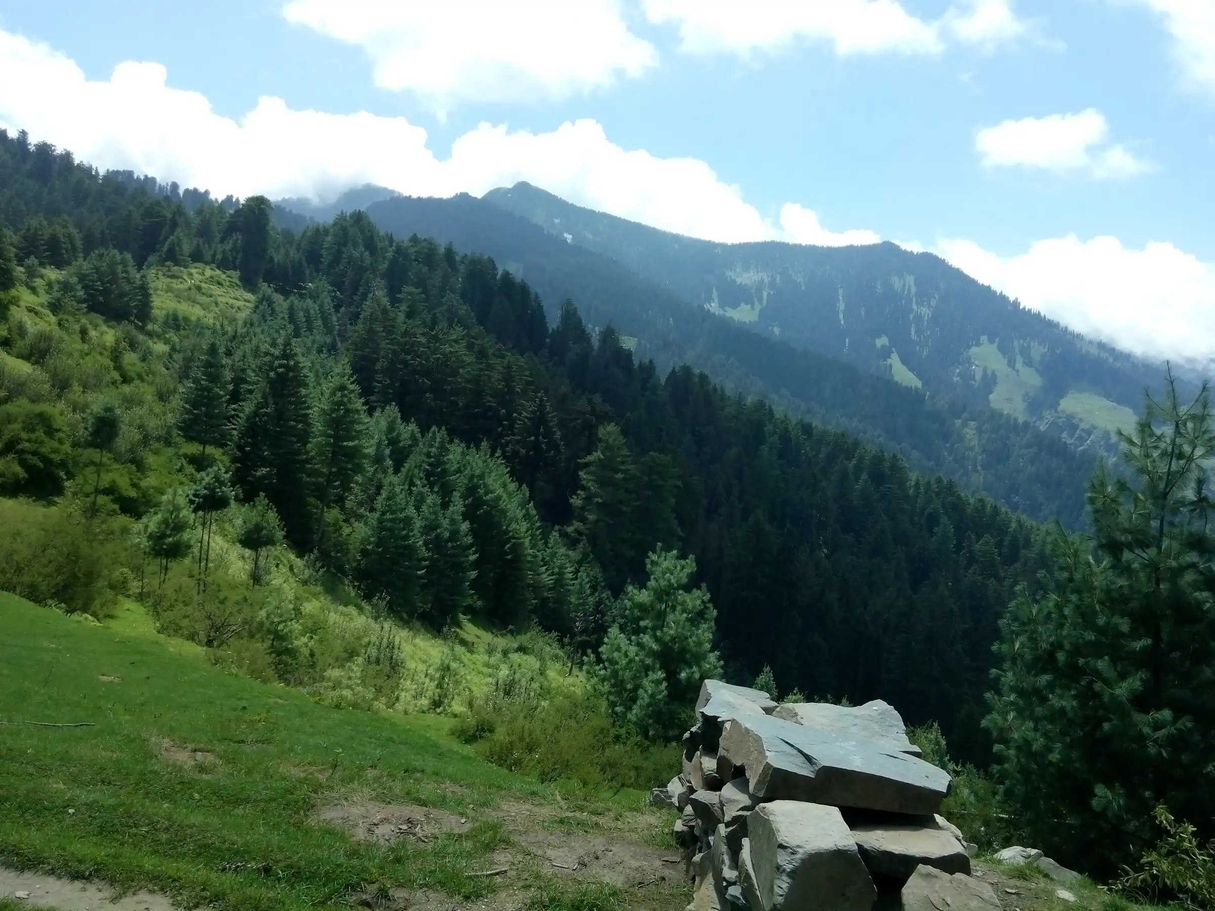 Forested slopes and a green valley seen from the Kais Dhar trail, with a stack of slate stones in the foreground