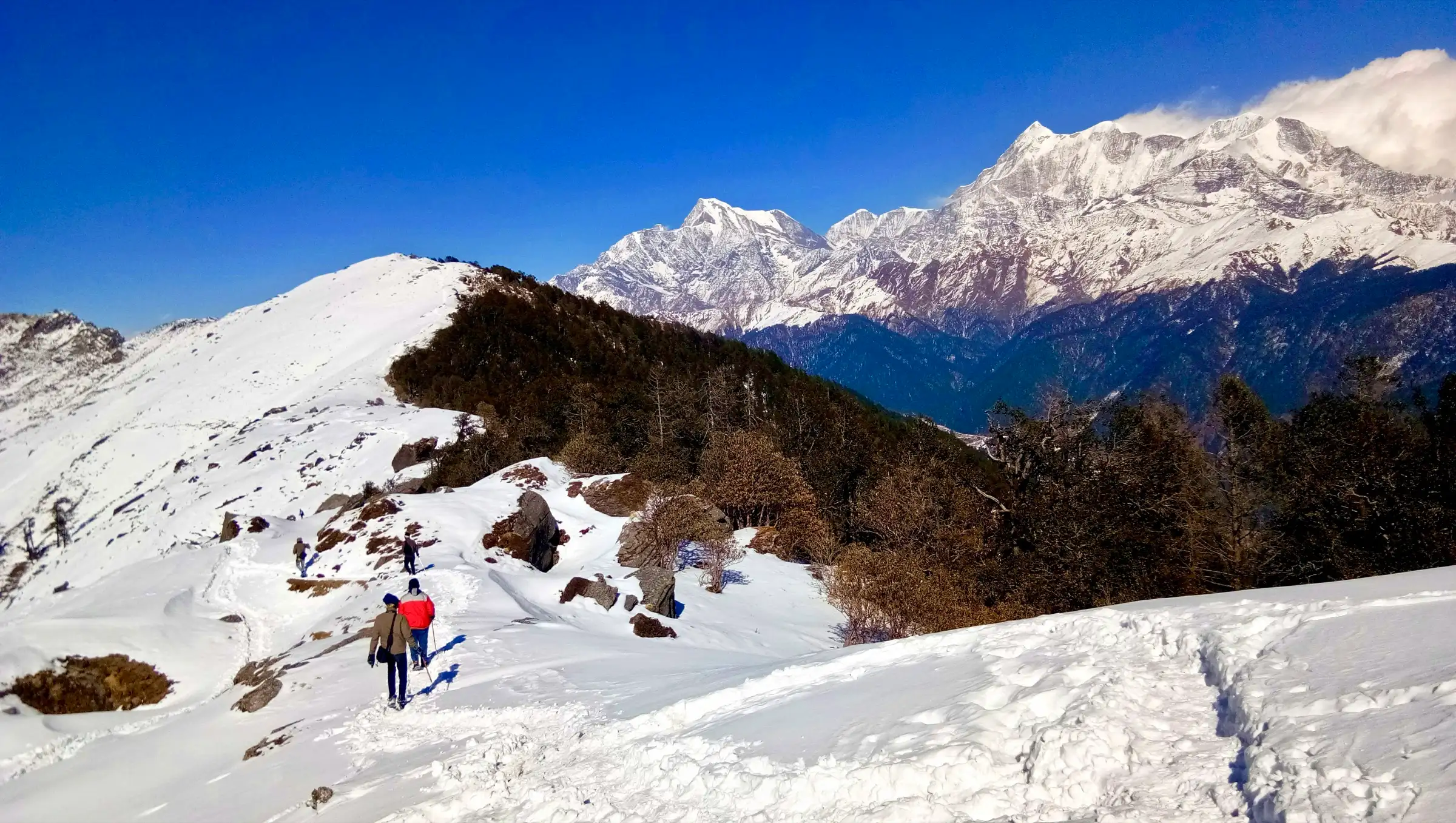 Trekkers walking a snowy ridge on the way to Brahmatal with the snow peaks of Trishul and Nanda Ghunti behind