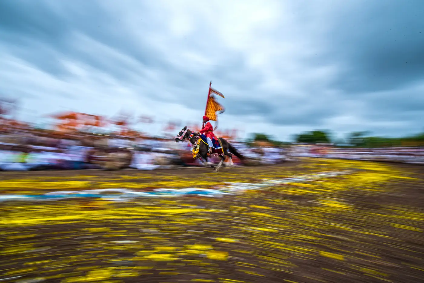 A rider in red with a saffron flag galloping round the flower-strewn ringan ground near Khudus Phata, Malshiras, under monsoon clouds