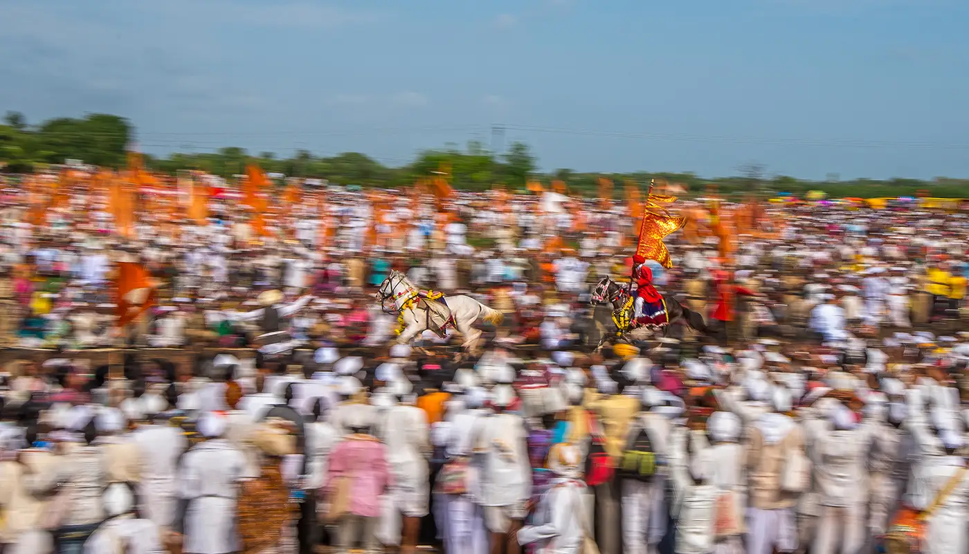 Two horses, one carrying a rider with a saffron flag, running the circular ringan through a vast crowd of warkaris near Velapur