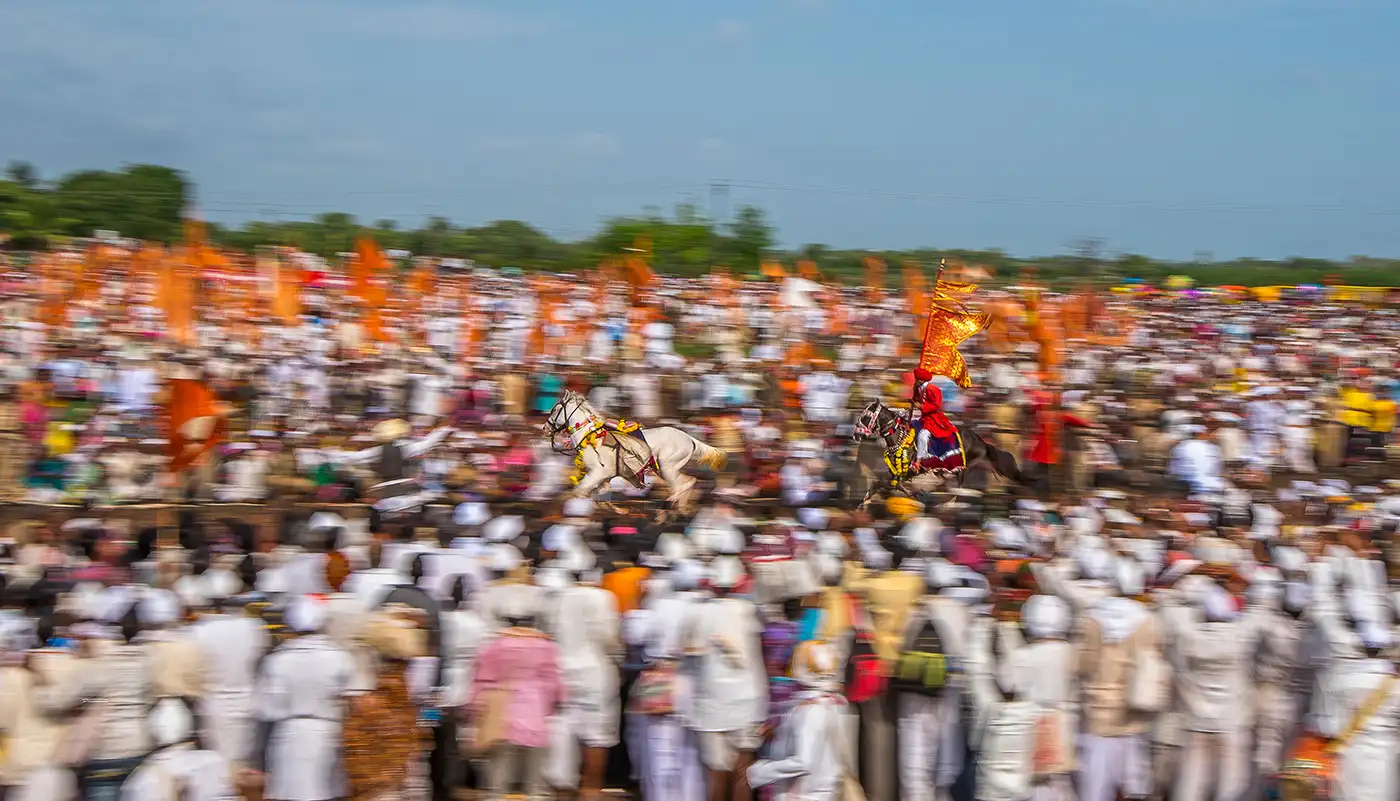 Two horses, one carrying a rider with a saffron flag, running the circular ringan through a vast crowd of warkaris near Velapur