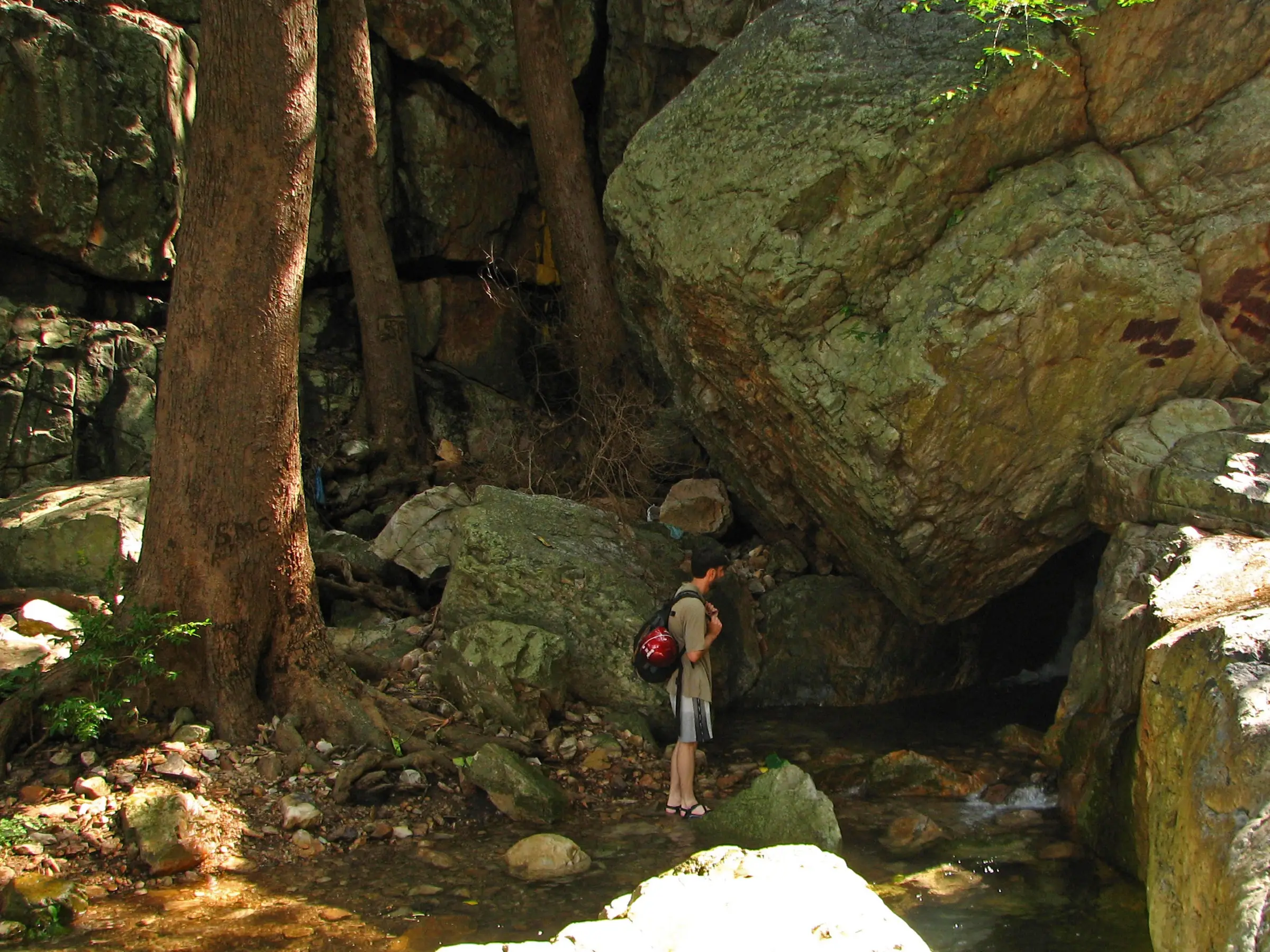 A trekker with a red backpack stands in the shallow stream below giant boulders in the narrowing Tada canyon