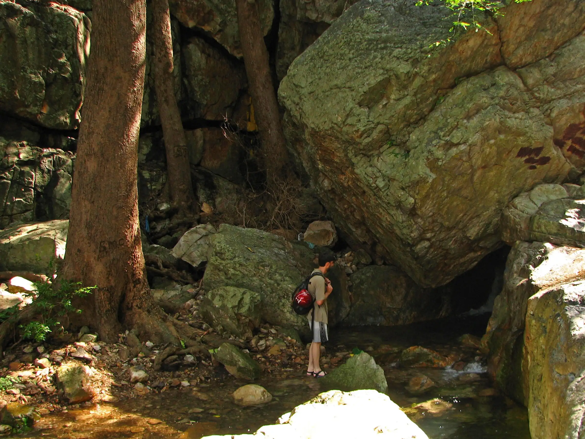 A trekker with a red backpack stands in the shallow stream below giant boulders in the narrowing Tada canyon