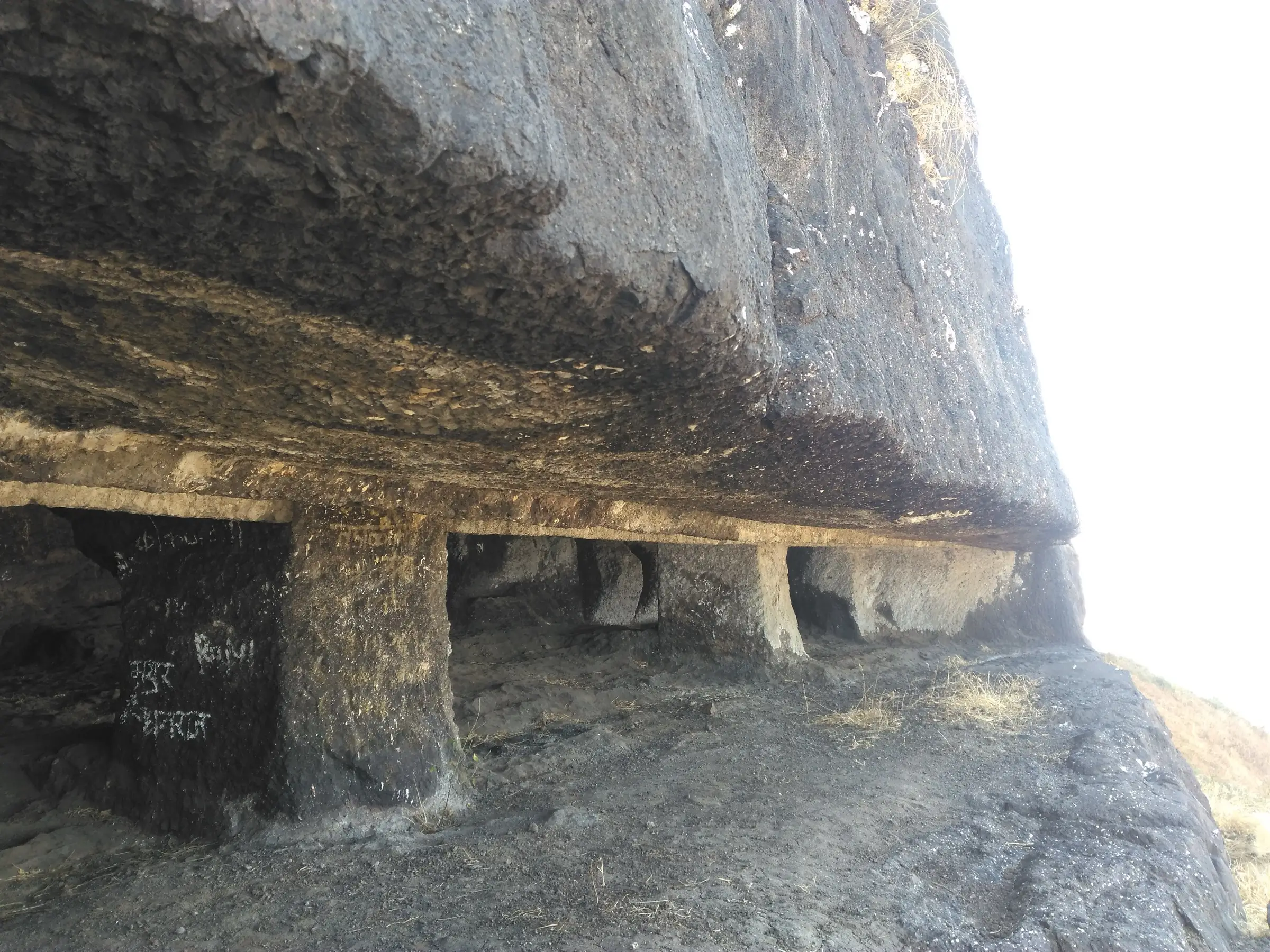 Pillared rock-cut caves carved under an overhanging basalt cliff on Dhodap Fort