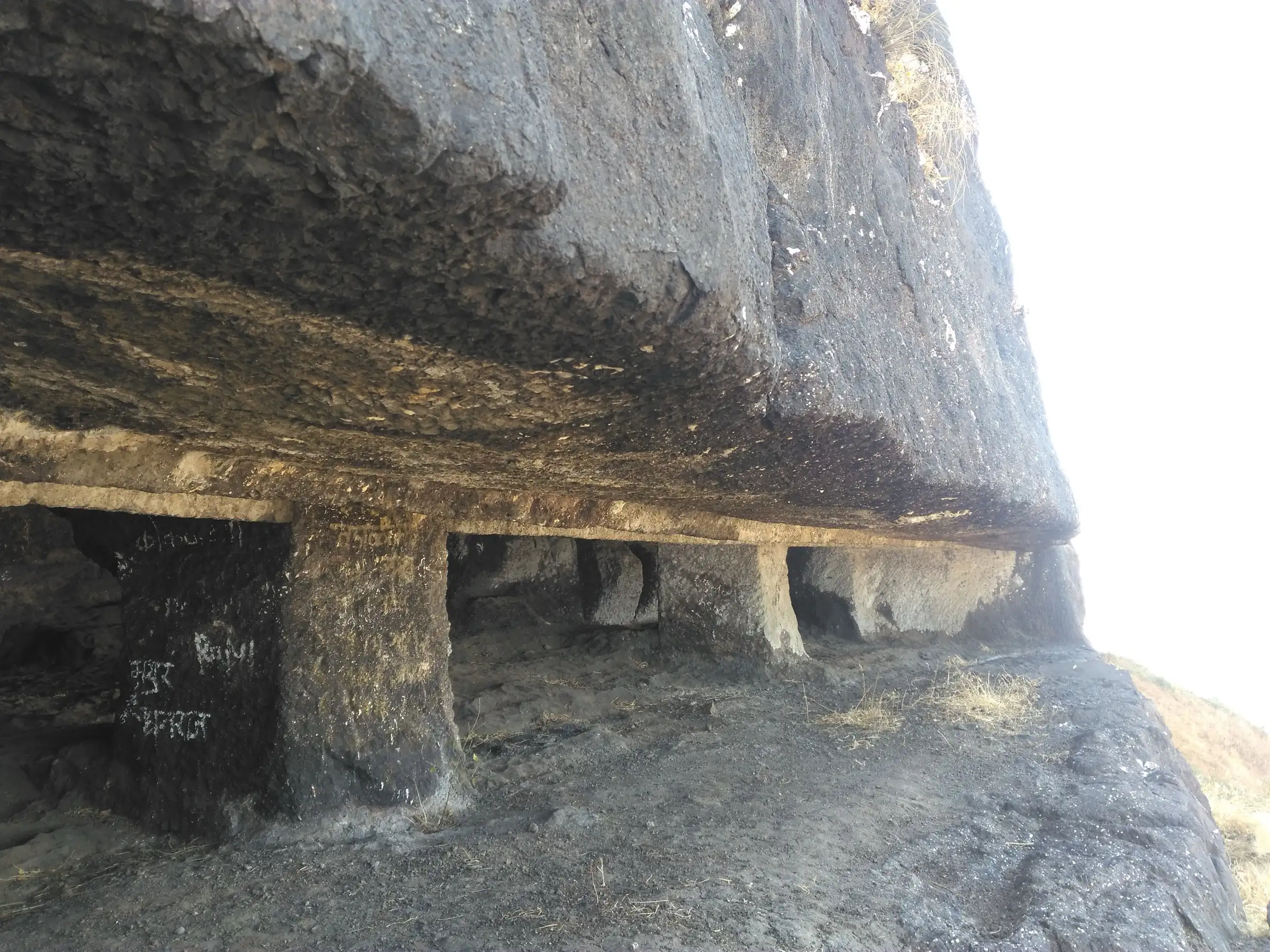 Pillared rock-cut caves carved under an overhanging basalt cliff on Dhodap Fort