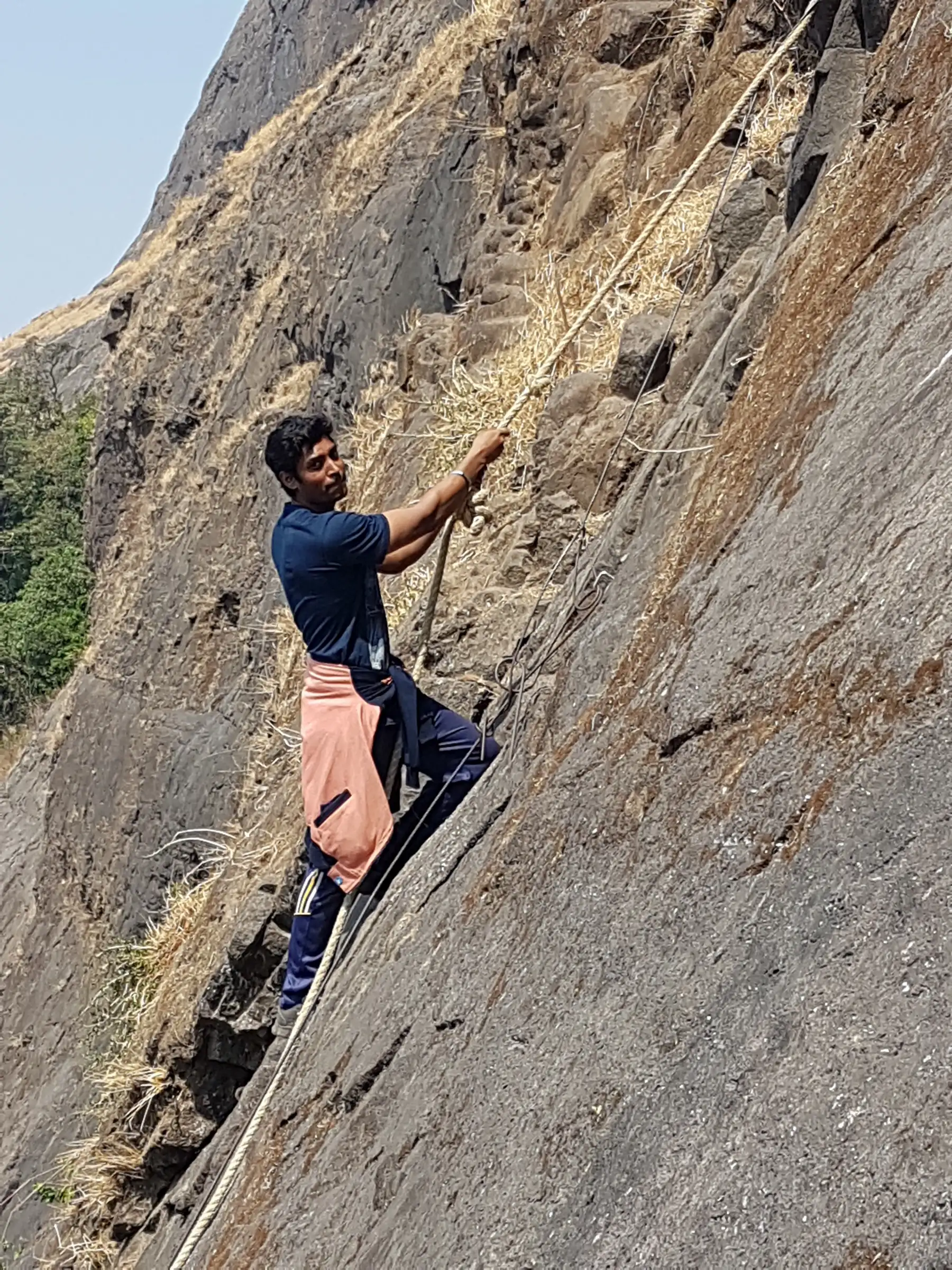 A trekker climbing a steep basalt slab holding a fixed rope on the way to the Dhak Bahiri cave