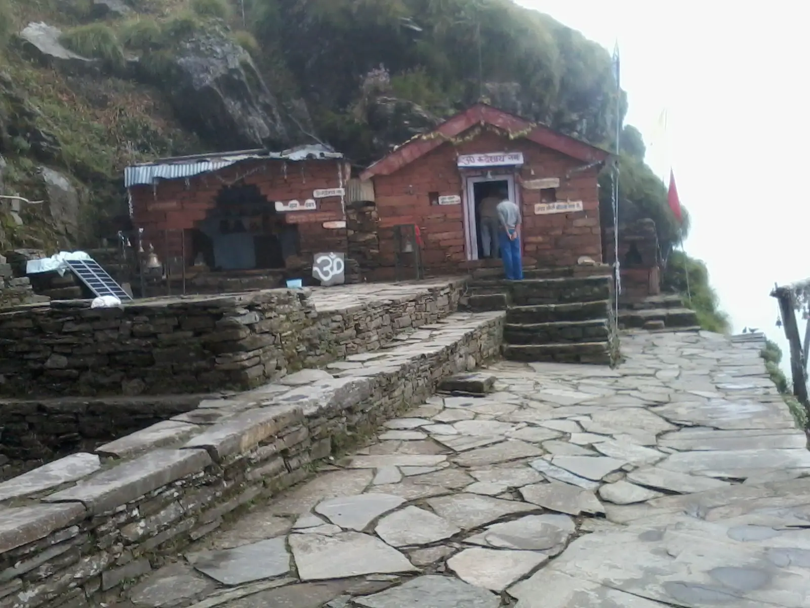 Small stone and brick shrines of Rudranath built into a mountain rock face, reached by a flagstone path in the mist
