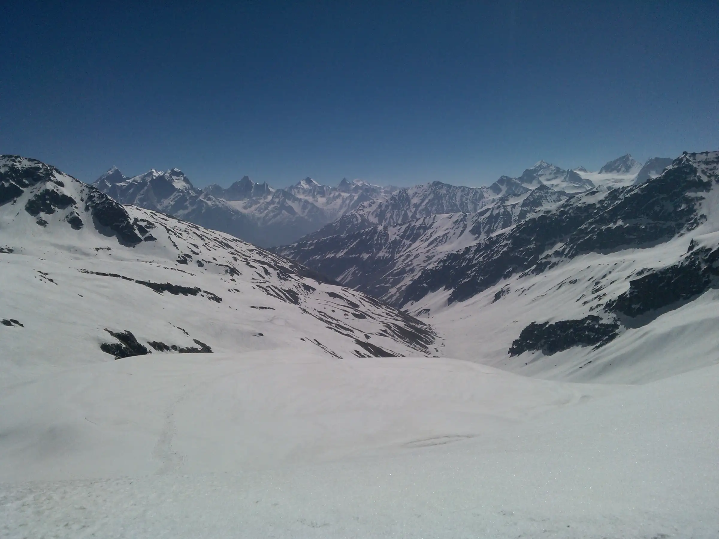 Snowfield on Rupin Pass looking down a snowy valley towards the jagged Kinner Kailash peaks under a deep blue sky