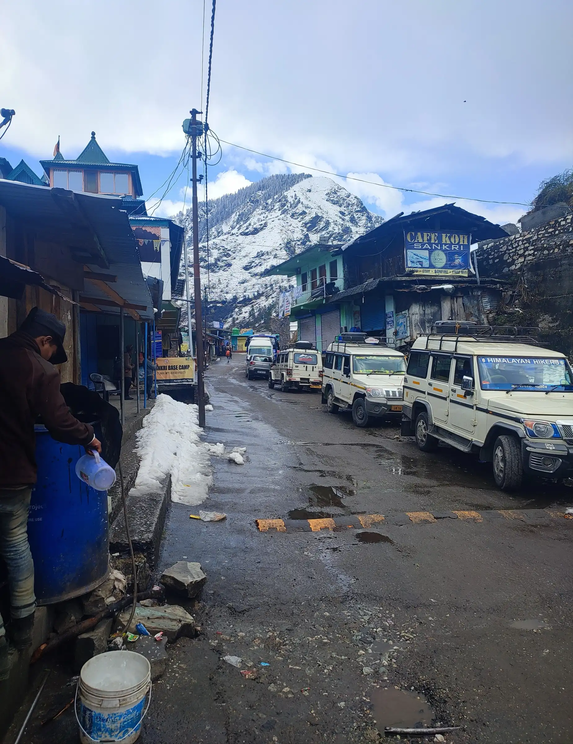 The main street of Sankri after snowfall, with shops, parked jeeps and a snow-covered peak behind