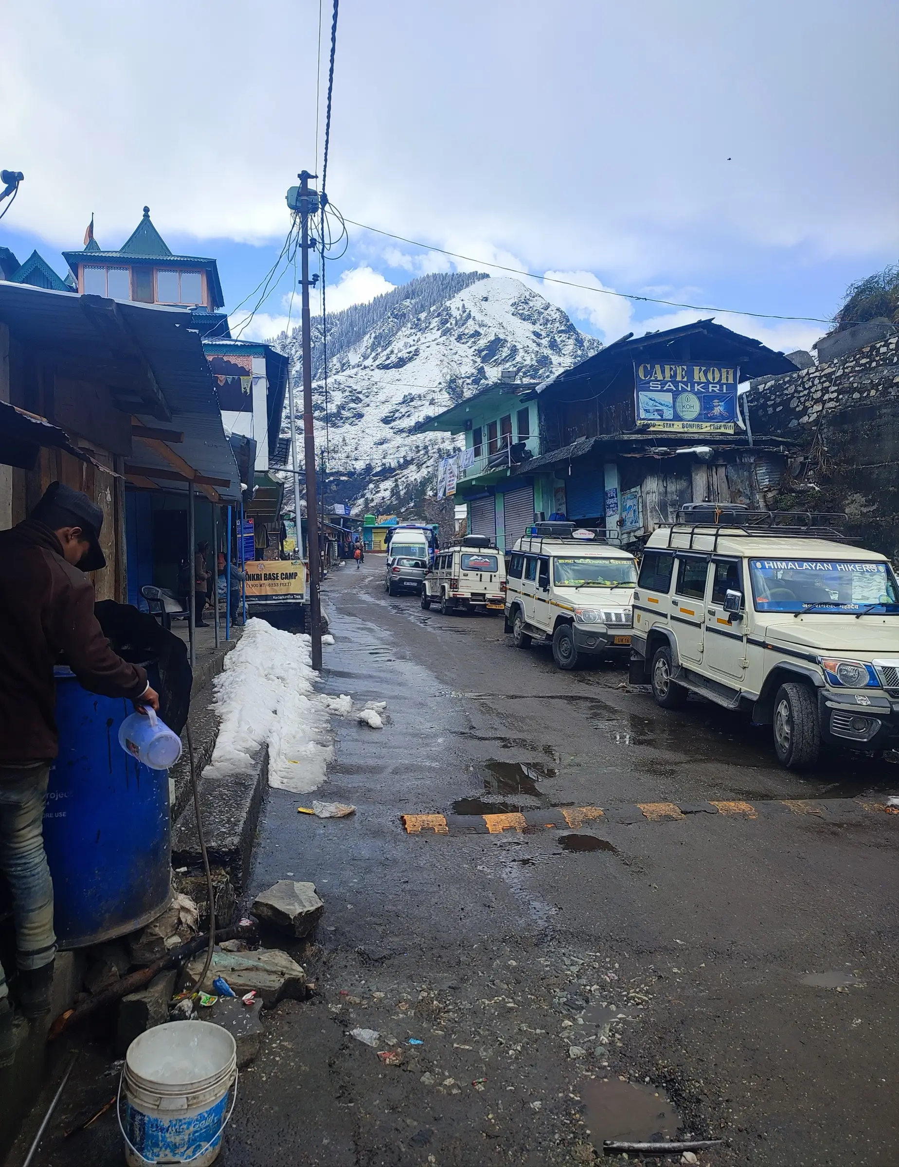 The main street of Sankri after snowfall, with shops, parked jeeps and a snow-covered peak behind