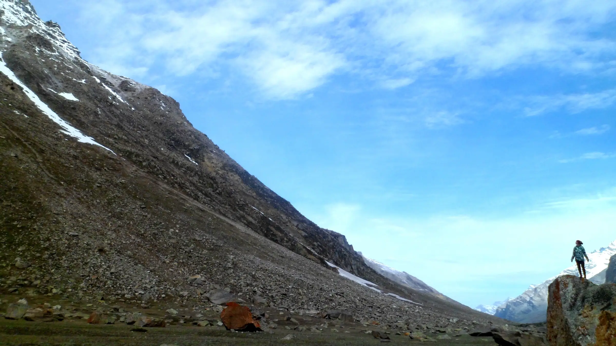 A trekker on a boulder beside a huge scree slope in the upper Parvati valley near Mantalai, under a blue sky