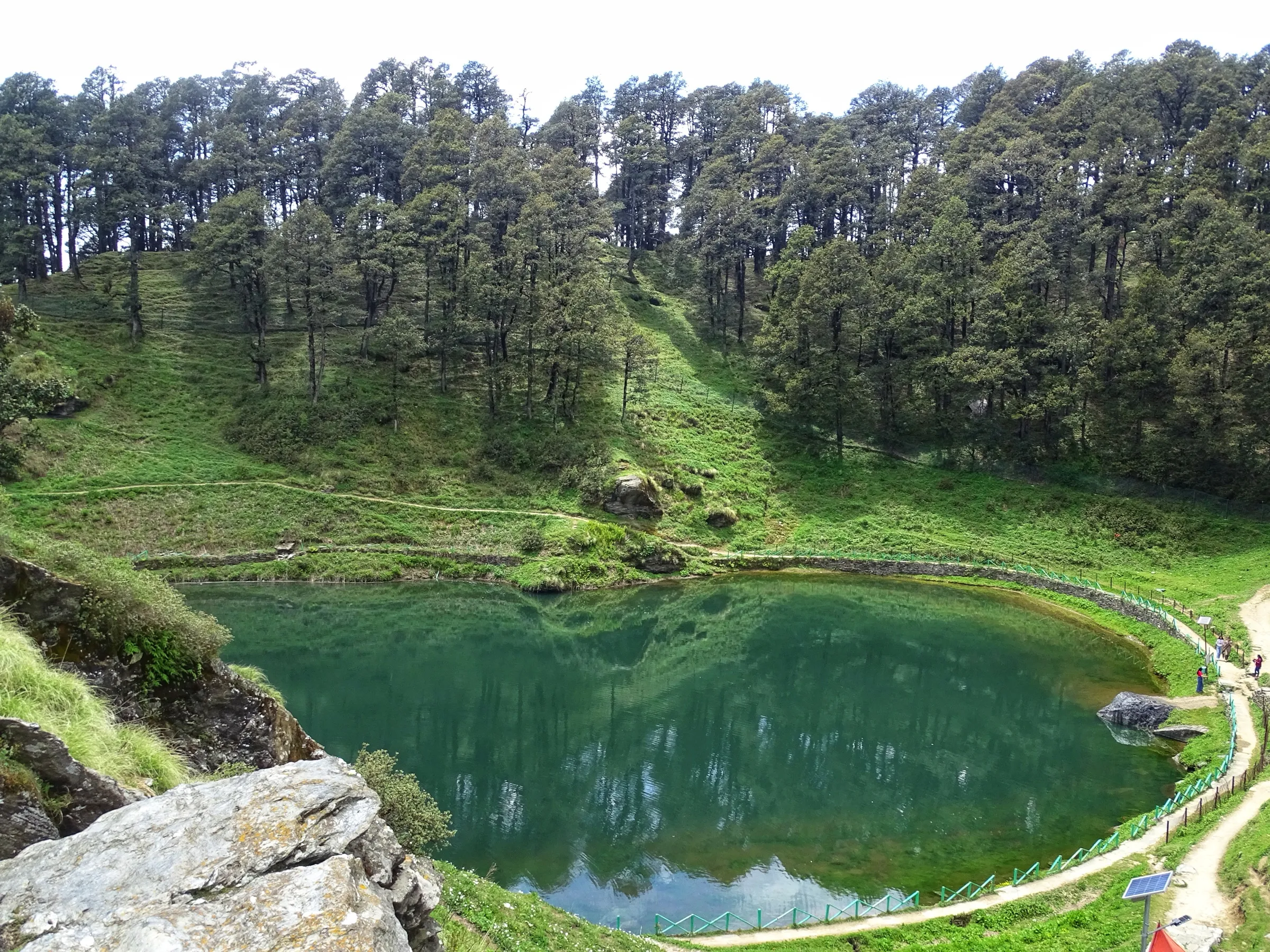Green Serolsar Lake seen from the rocks above, with a fenced path round the shore and tall conifers on the hillside behind