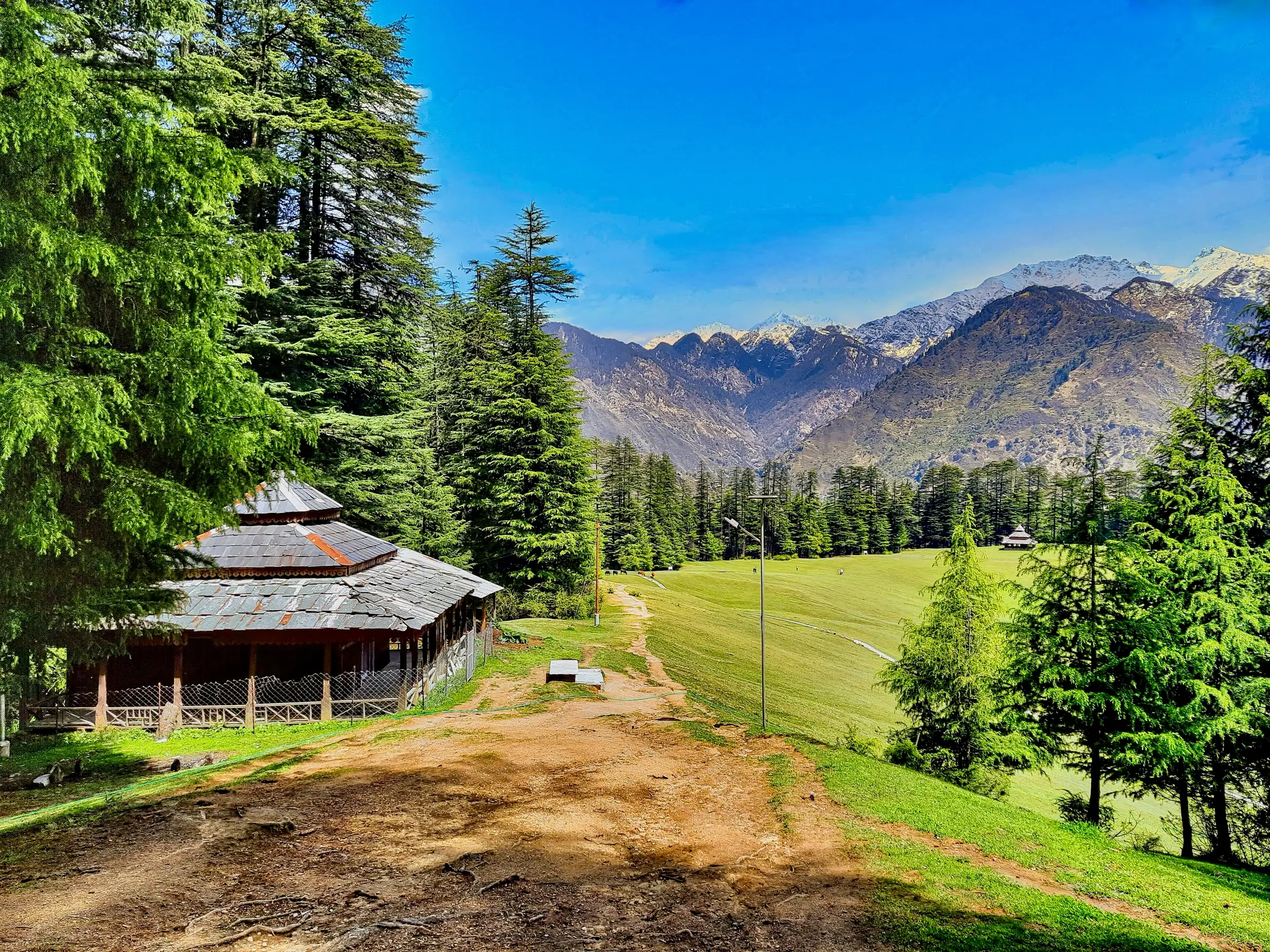 Path along the edge of the Shangarh meadow past a wooden temple building, with snow peaks of the Sainj valley behind
