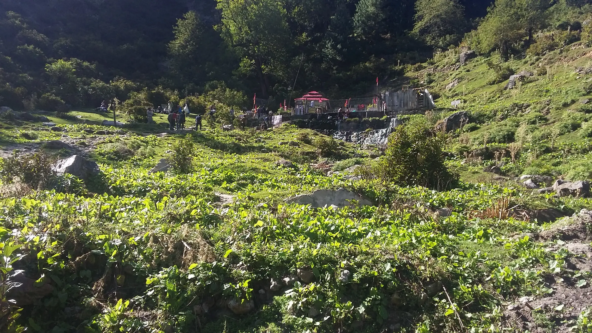 Green hillside at Kheerganga leading up to the Shiv temple and the hot-spring bathing tank, with pilgrims beside the stream