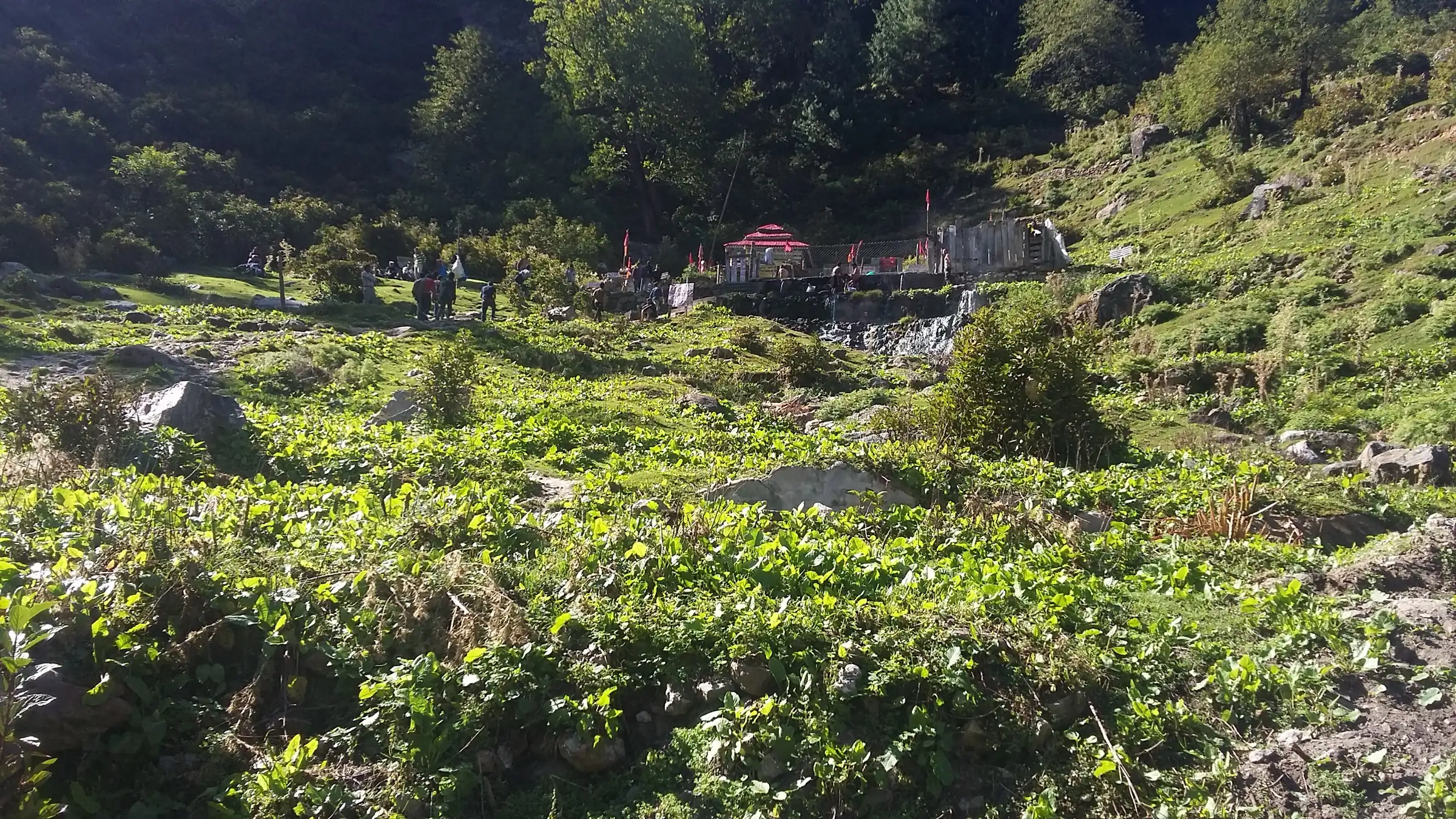 Green hillside at Kheerganga leading up to the Shiv temple and the hot-spring bathing tank, with pilgrims beside the stream