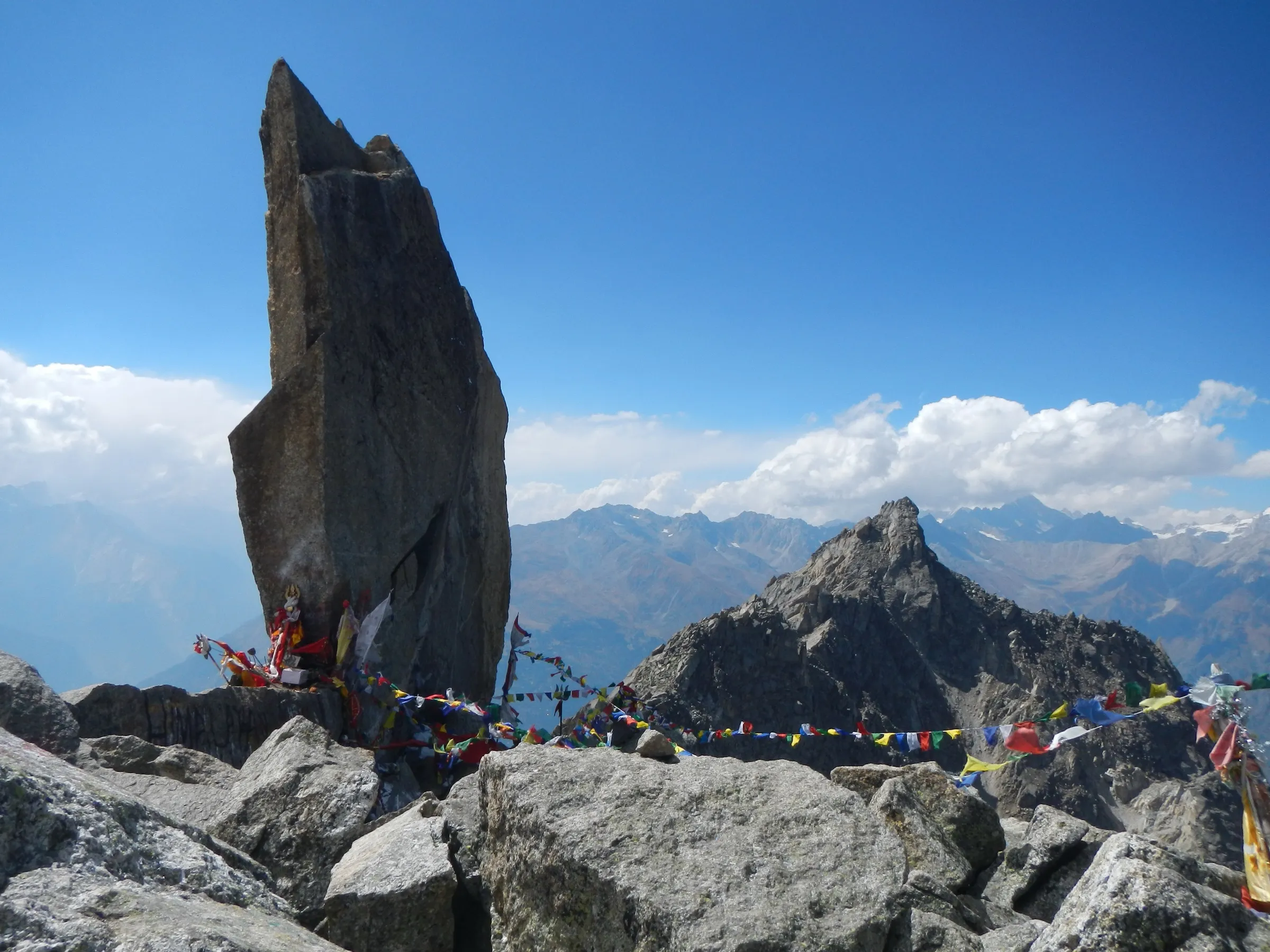 The tall Kinner Kailash Shivling rock pillar with prayer flags on a boulder ridge, mountains and clouds behind
