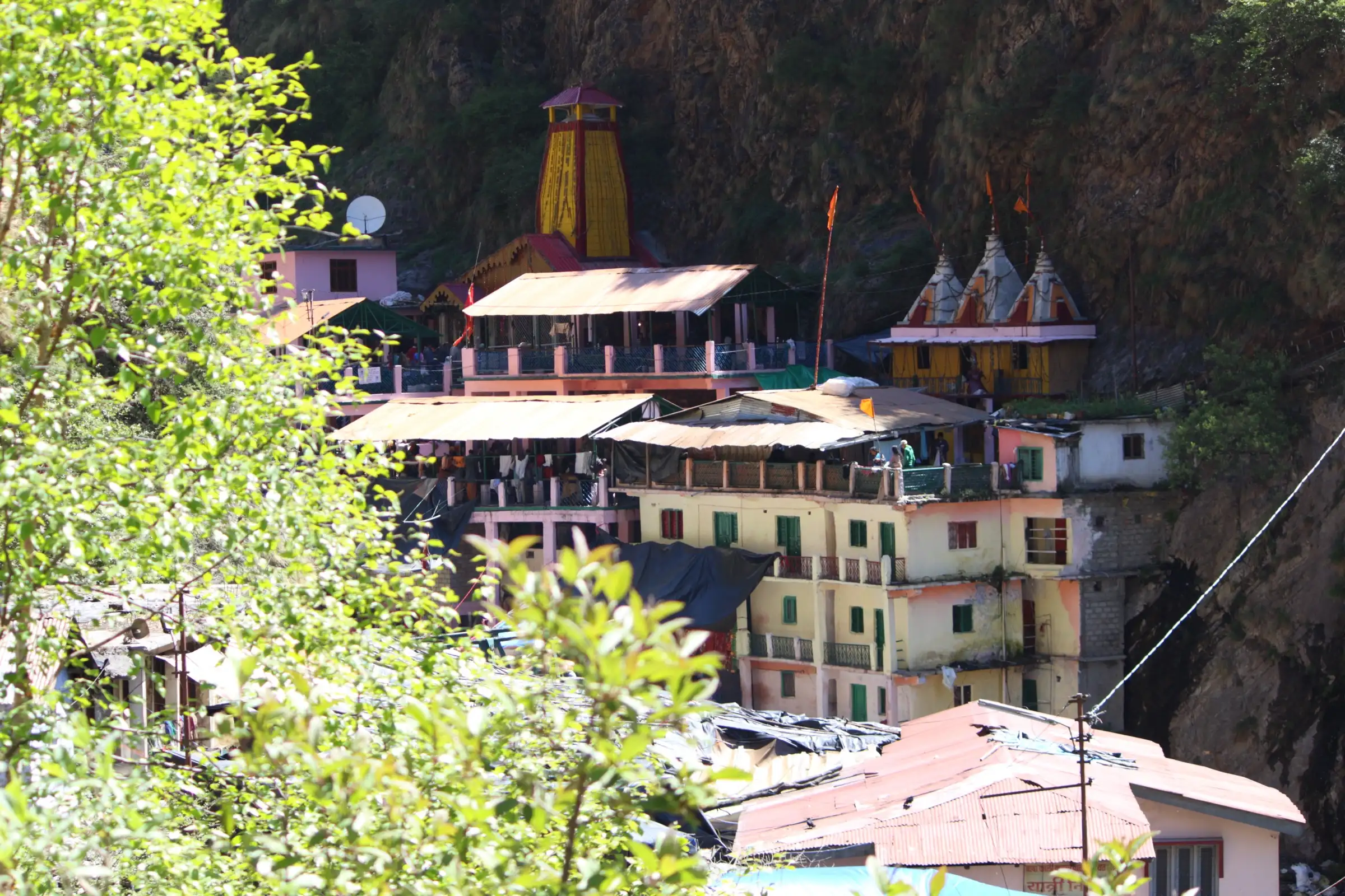 The yellow spire of the Yamunotri temple and pilgrim buildings against a dark cliff, seen through green leaves
